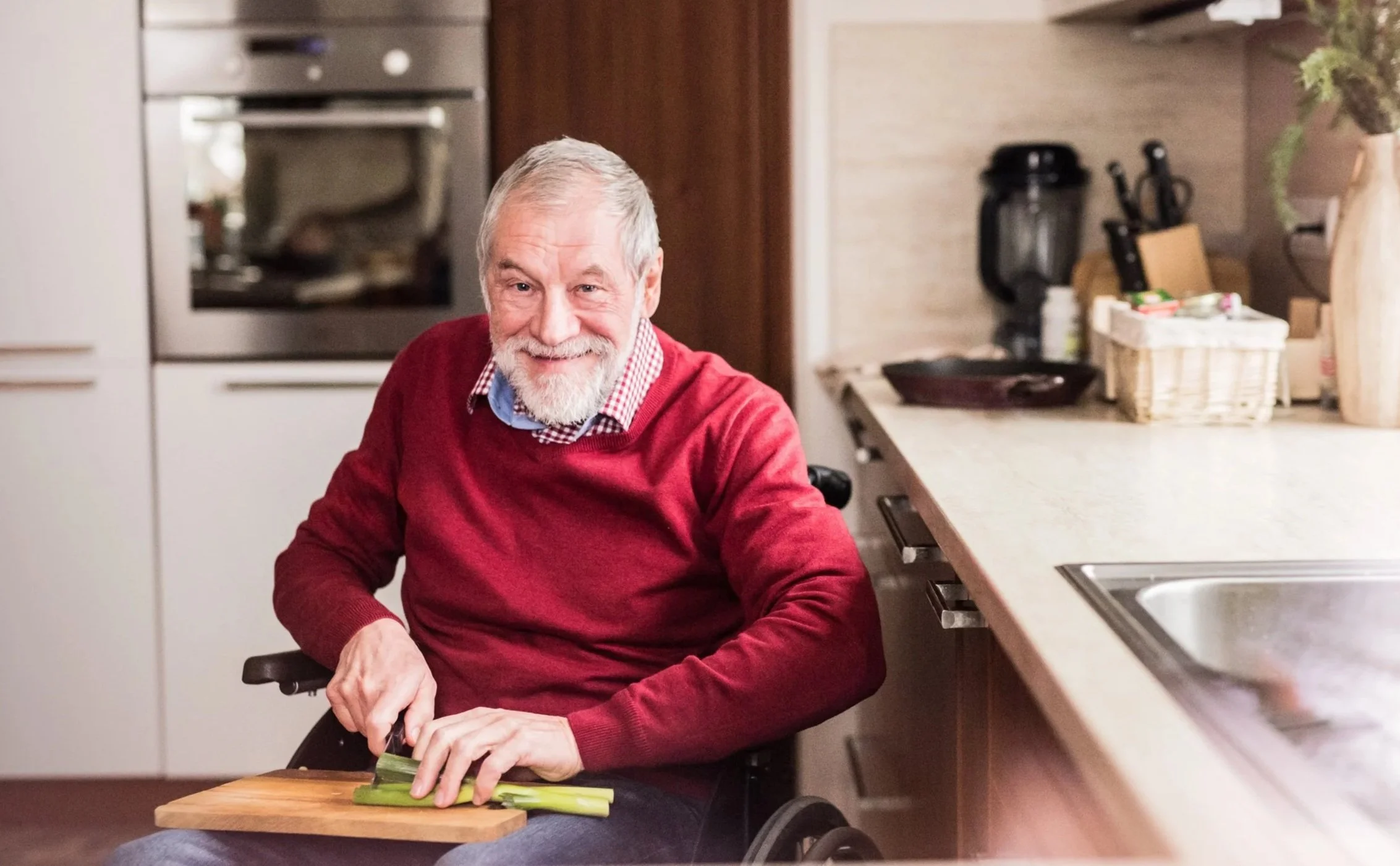 An elderly man with a white beard wearing a red sweater and checkered shirt, sitting in a wheelchair, chopping celery on a wooden cutting board in a kitchen.