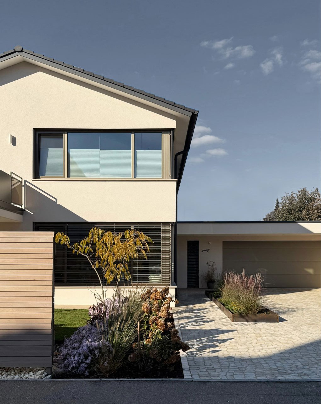 Moderne zweistöckige Hausfassade mit großen Fenstern, gepflegtem Vorgarten mit Pflanzen, Ziersträuchern und einer gepflasterten Einfahrt unter hellem Himmel.