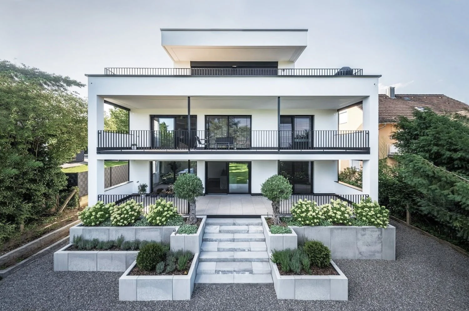 Modernes weißes Mehrfamilienhaus mit drei Stockwerken, umgeben von gepflegten Pflanzen, Bäumen und einem gepflasterten Vorgarten