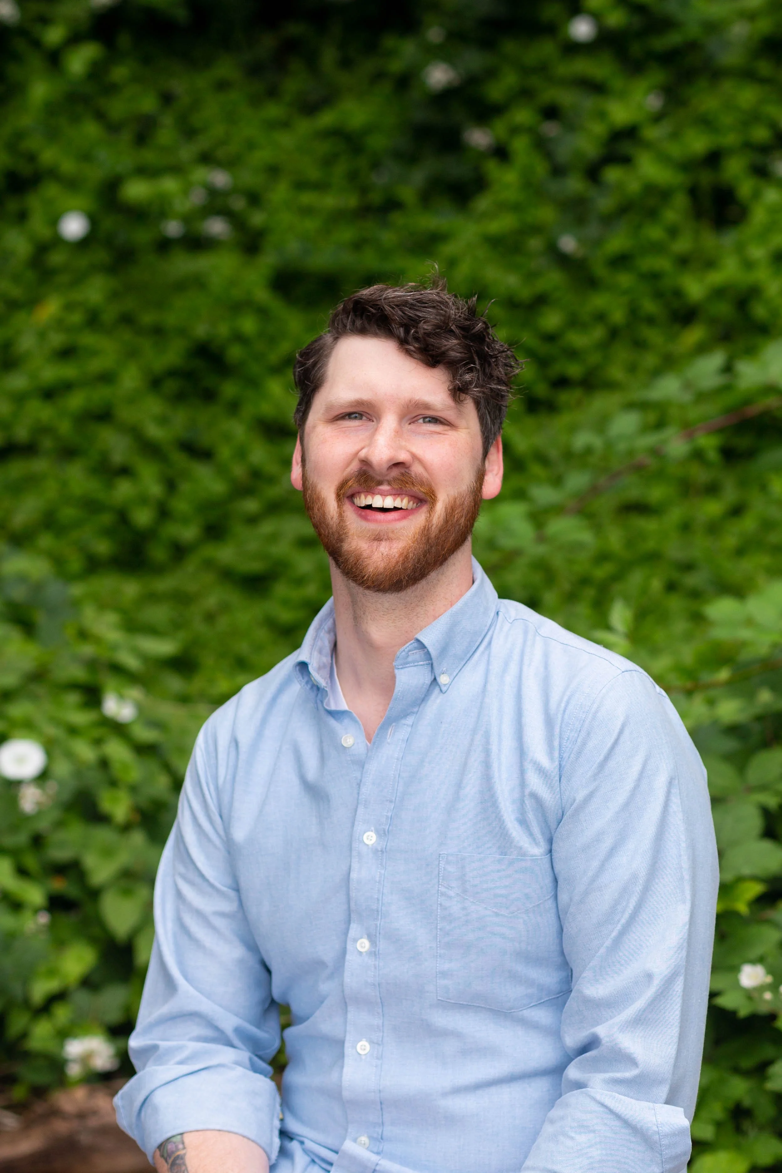 A smiling man with a beard and mustache, wearing a light blue button-up shirt, sitting outdoors in front of green foliage.