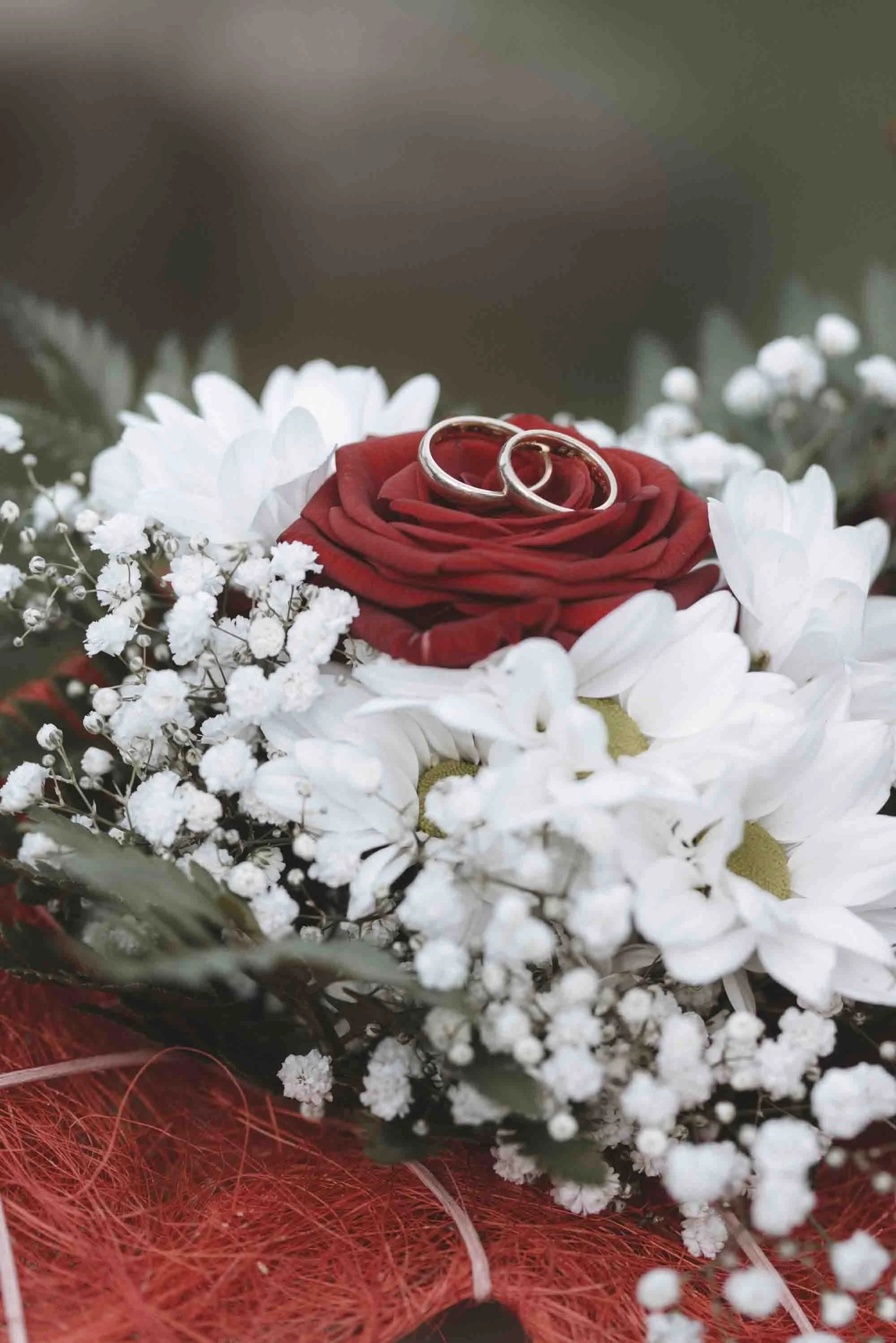 Zwei Eheringe liegen auf einer roten Rose, die auf einer Blumenstrauß mit weißen Blumen und kleinen weißen Beeren liegt.