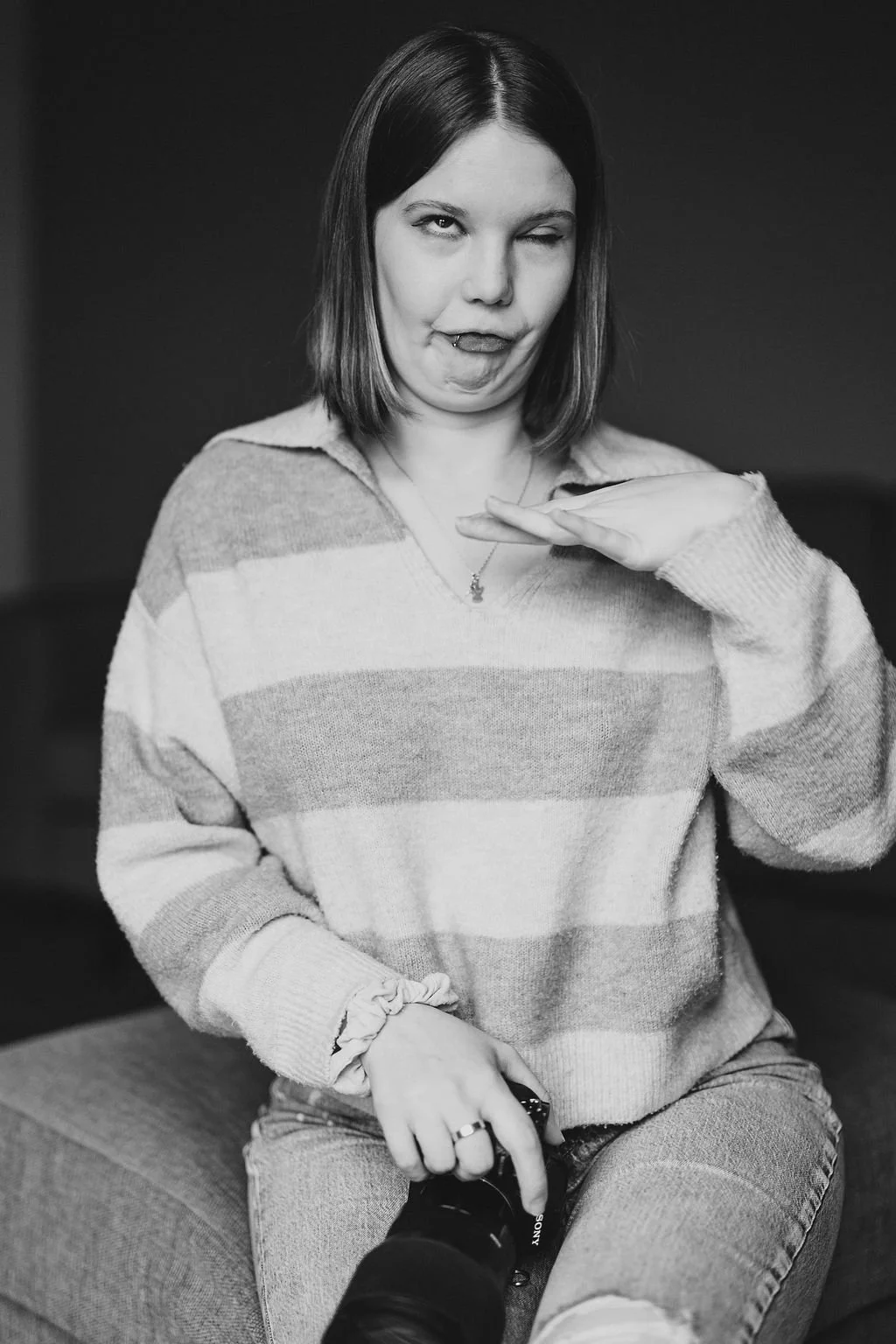 Schwarz-weiß Foto einer jungen Frau mit Schulterlänge Haaren, die eine lustige Grimasse macht, während sie mit der Hand eine Pose andeutet. Sie trägt einen gestreiften Pullover und sitzt auf einem Hocker.