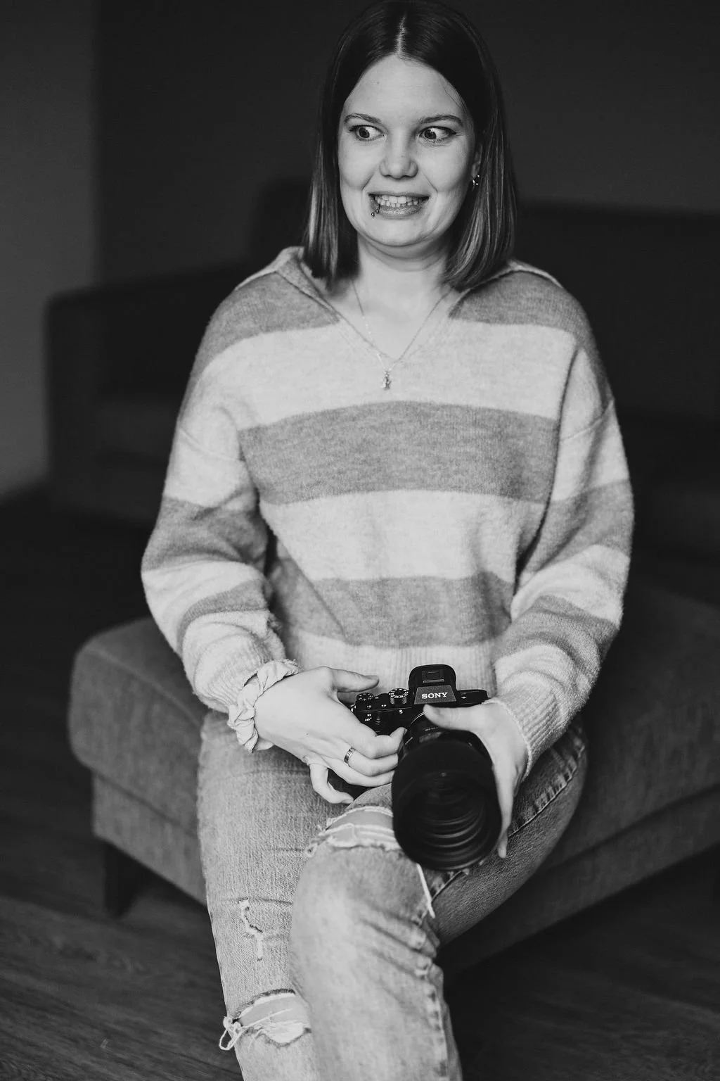 Junge Frau sitzt auf einem Sofa, hält eine Kamera in den Händen, trägt ein gestreiftes Pullover, Jeans mit Rissen. Das Bild ist schwarz-weiß.