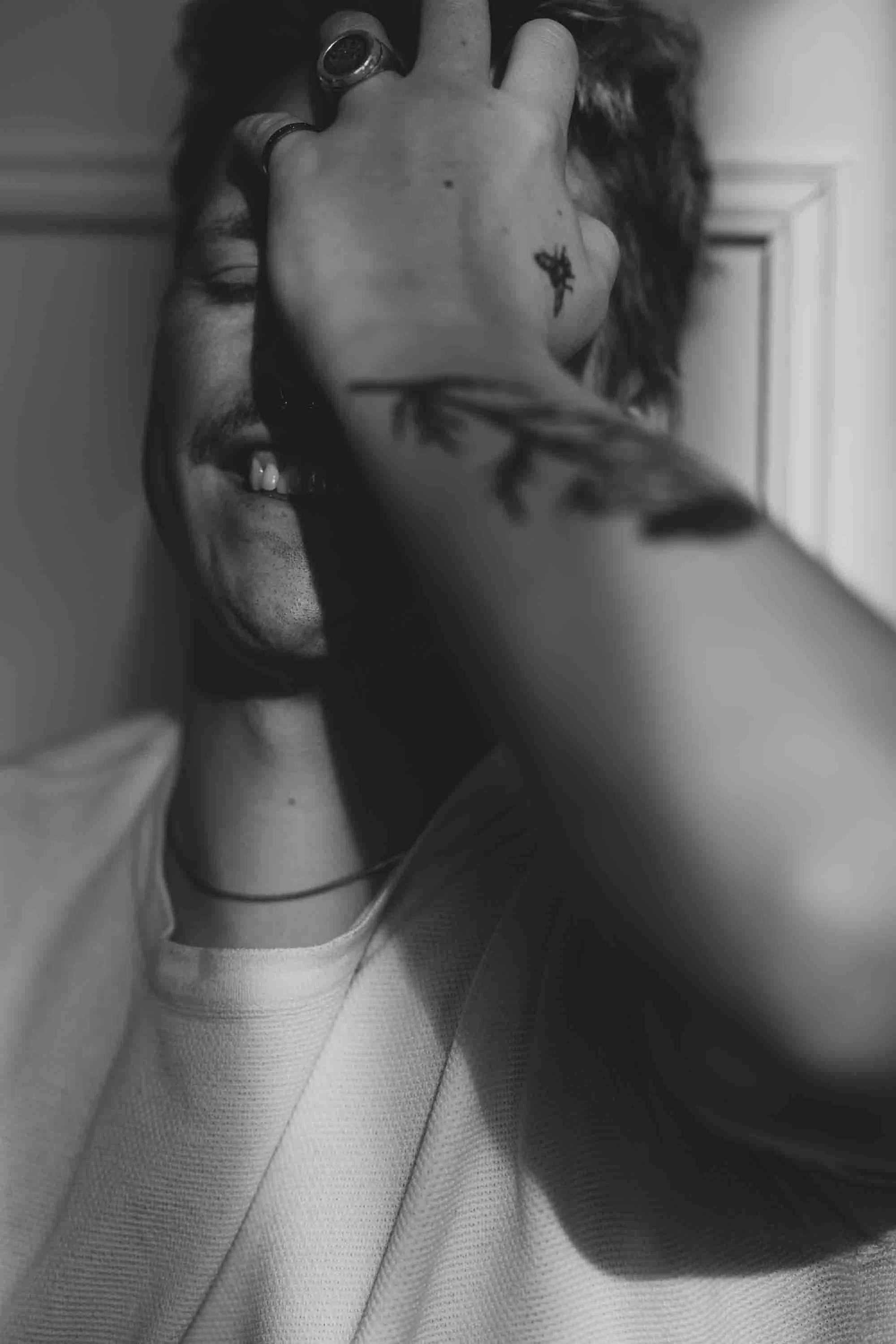 Schwarz-Weiß-Foto eines lachenden Mannes, der eine Hand vor das Gesicht hält, Blumentattoos sichtbar auf dem Arm, trägt Ringe und eine Halskette.
