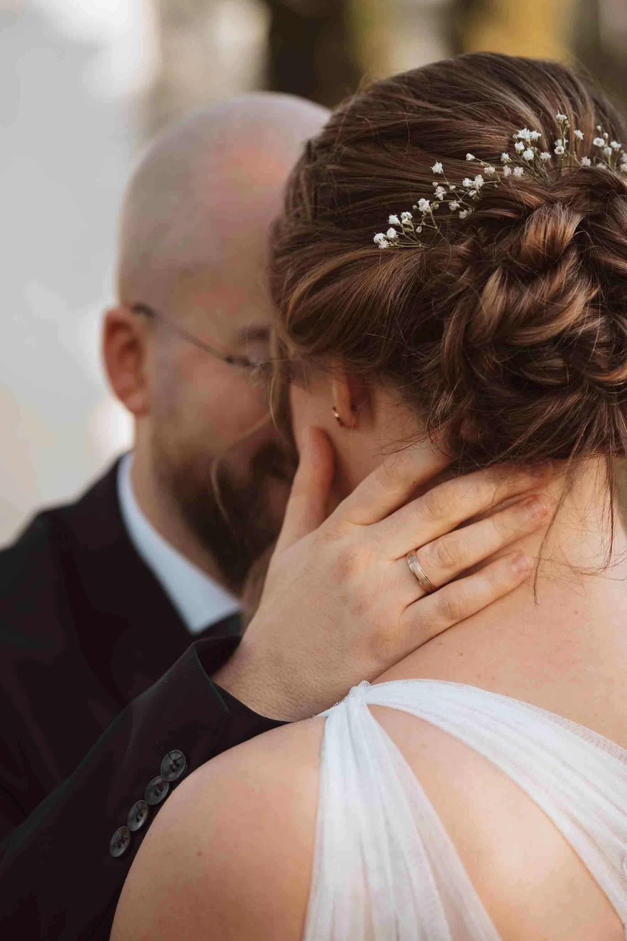 Ein Paar während einer Hochzeitszeremonie, das sich küssend berührt, eine Frau mit geflochtener Brautfrisur und weißen Blumen, ein Mann in einem dunklen Anzug.