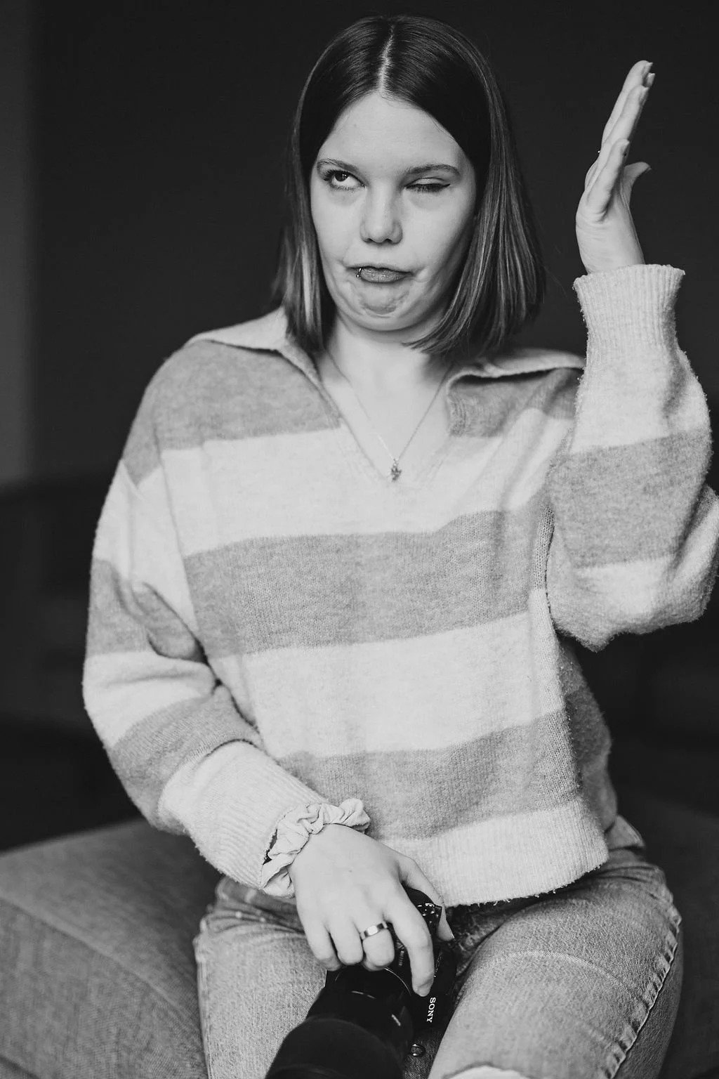 Schwarz-weiß-Foto einer jungen Frau mit kurzen braunen Haaren, die eine Strickpullover trägt, einen Kamera in der Hand hält und beim Blick in die Kamera eine fragende oder schmunzelnde Mimik zeigt.