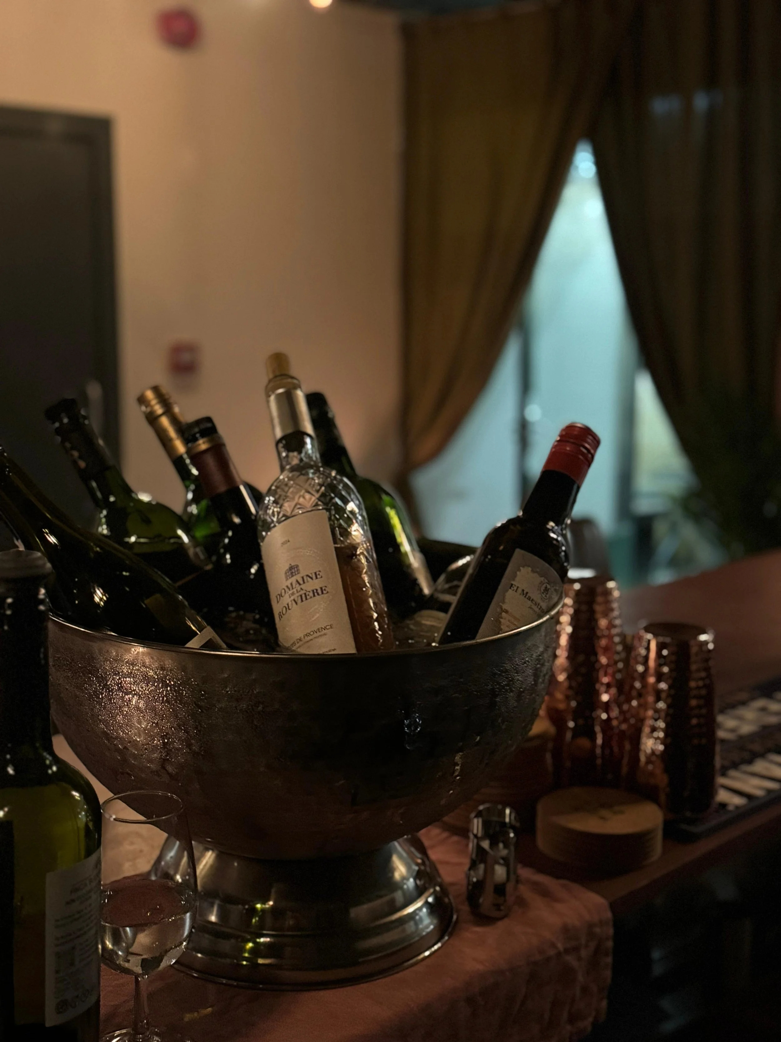 A large bowl filled with various bottles of wine and a glass of water on a table, with a stack of cups and a curtain-covered window in the background.