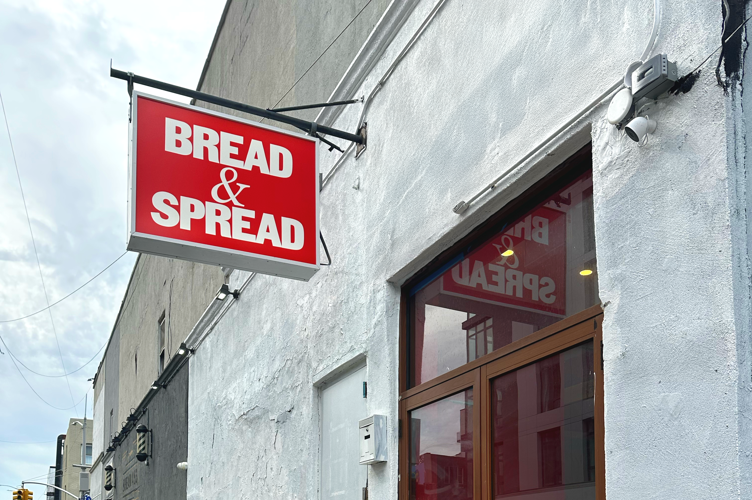Red and white sign reading 'Bread & Spread' attached to a white exterior wall of a building. Reflects in the large window below the sign.