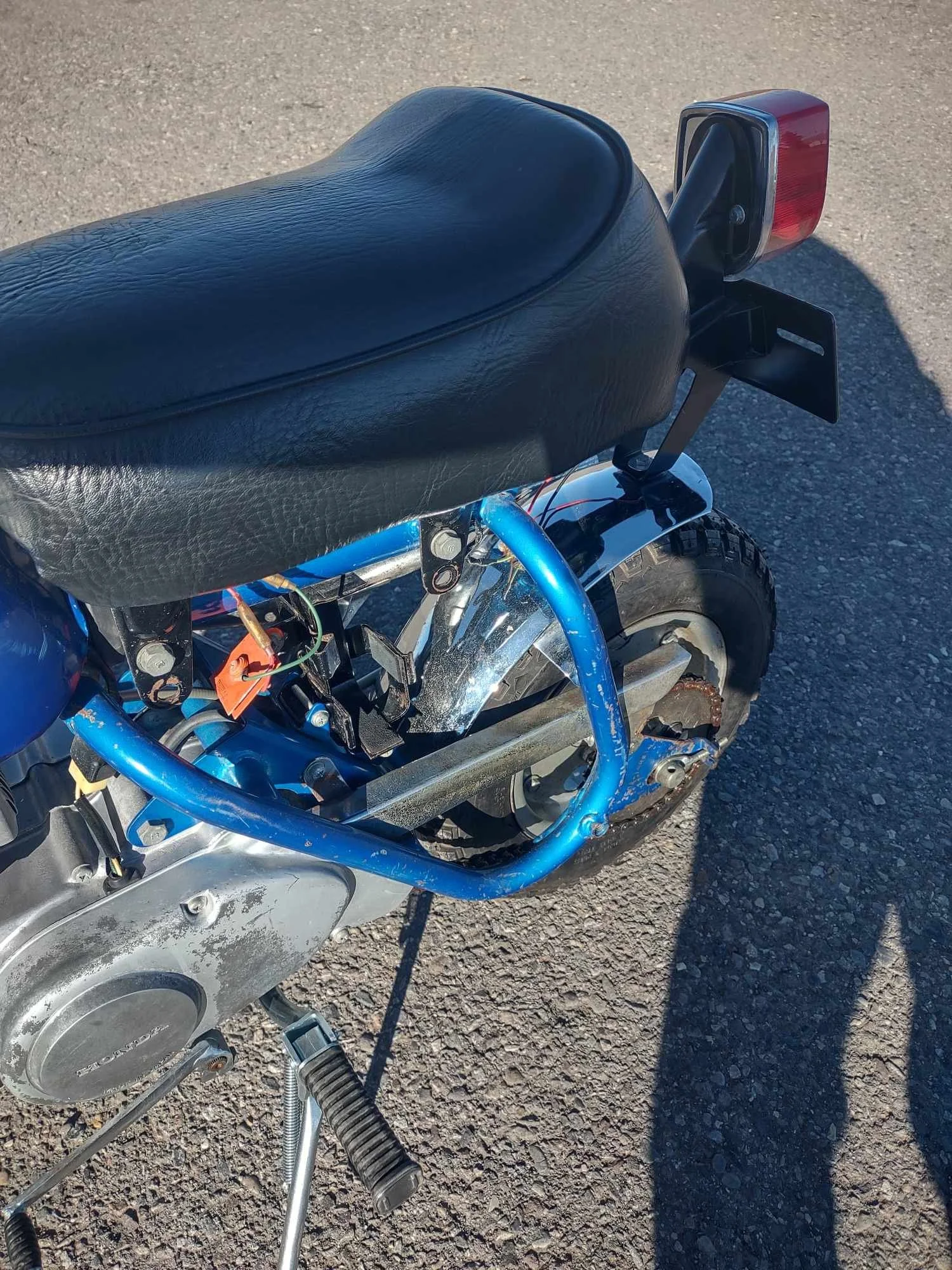 Close-up of the rear and left side of a small, blue scooter with a black seat, red tail light, and exposed wires.