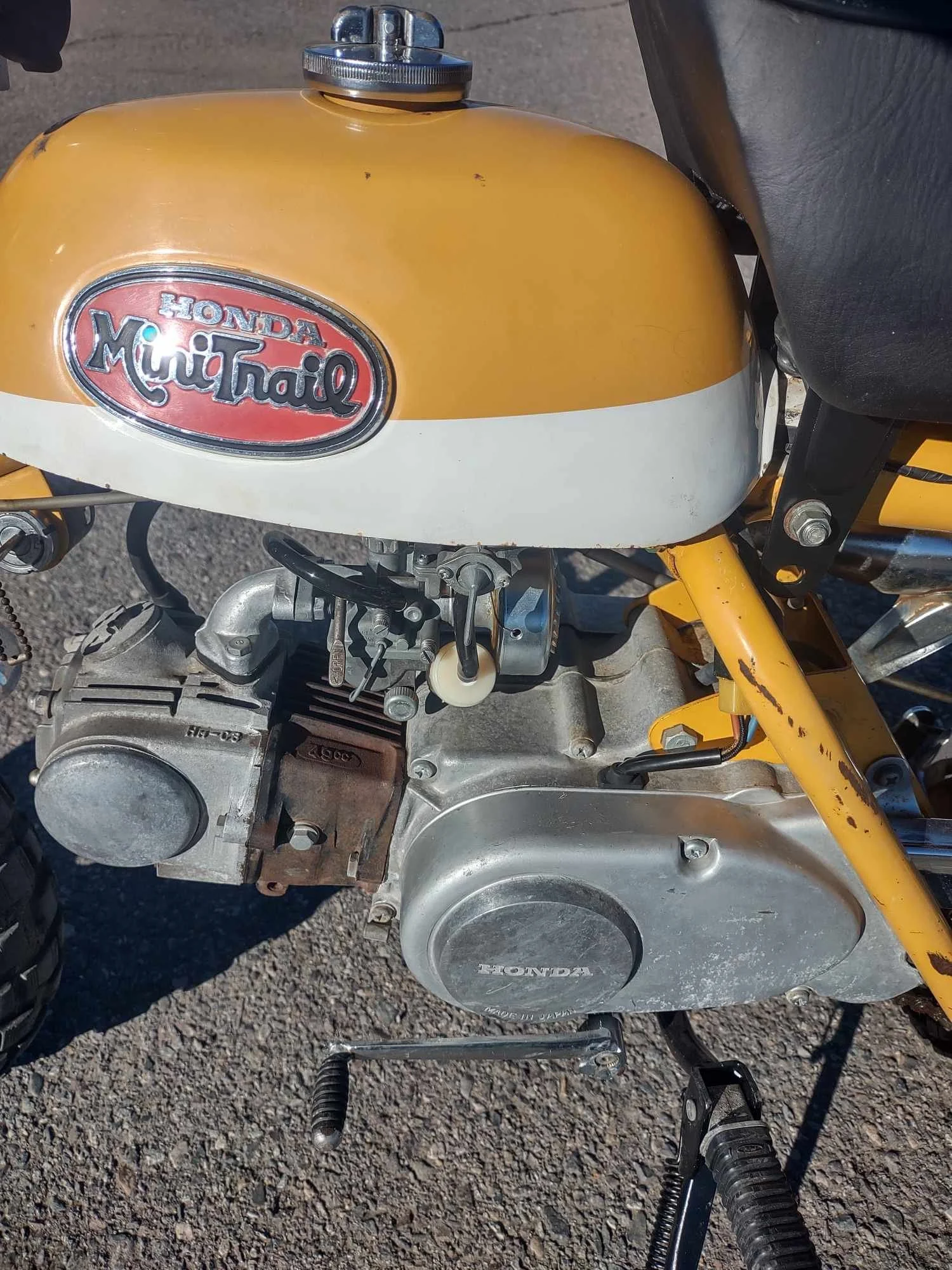Close-up of a vintage yellow Honda Mini Trail dirt bike with a silver engine labeled Honda. The bike's fuel tank features a red and silver Honda Mini Trail emblem. The black seat and yellow frame are visible.