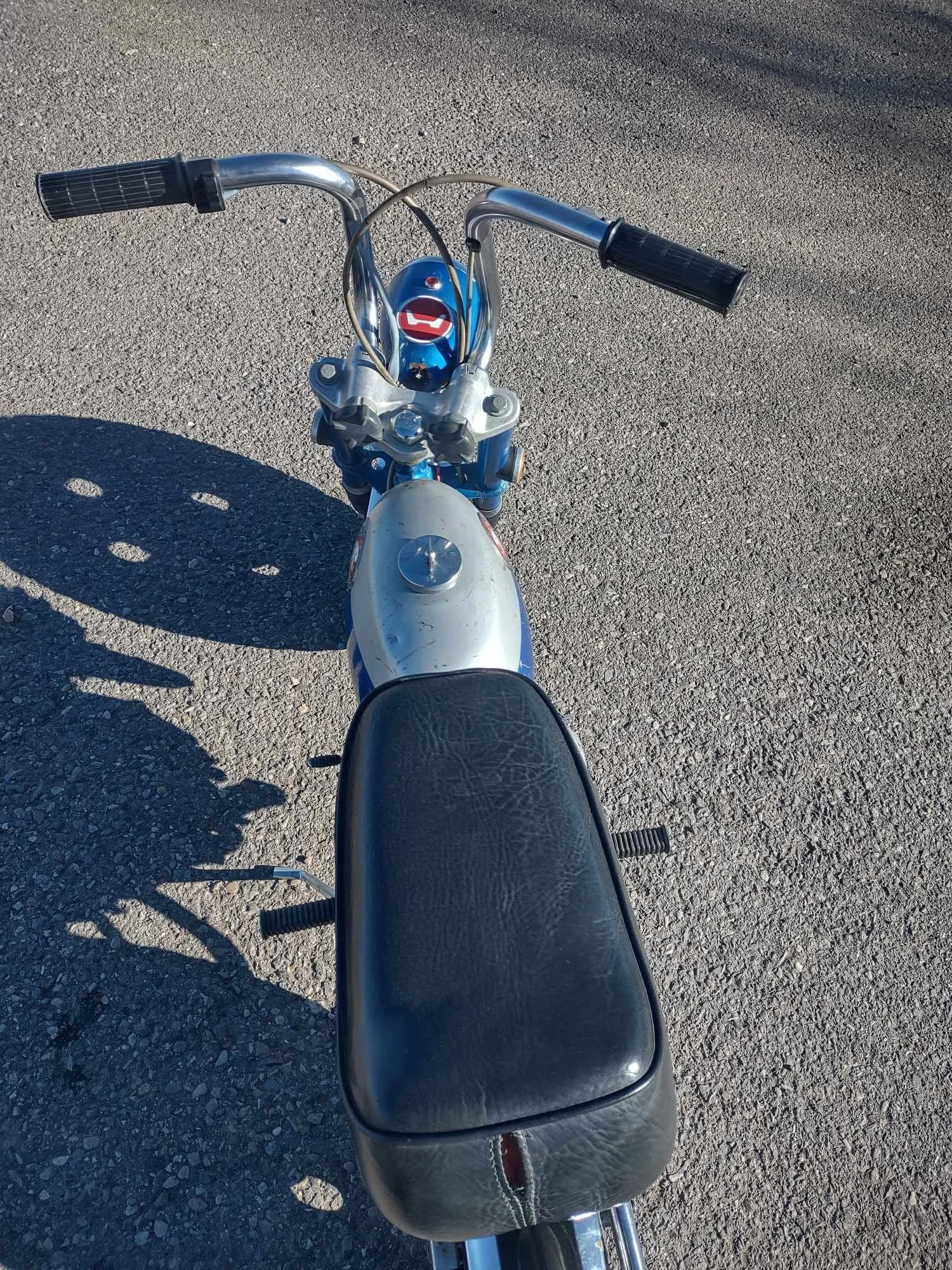 Top-down view of a vintage motorcycle with a black leather seat, chrome handlebars, and a speedometer, on a paved surface.