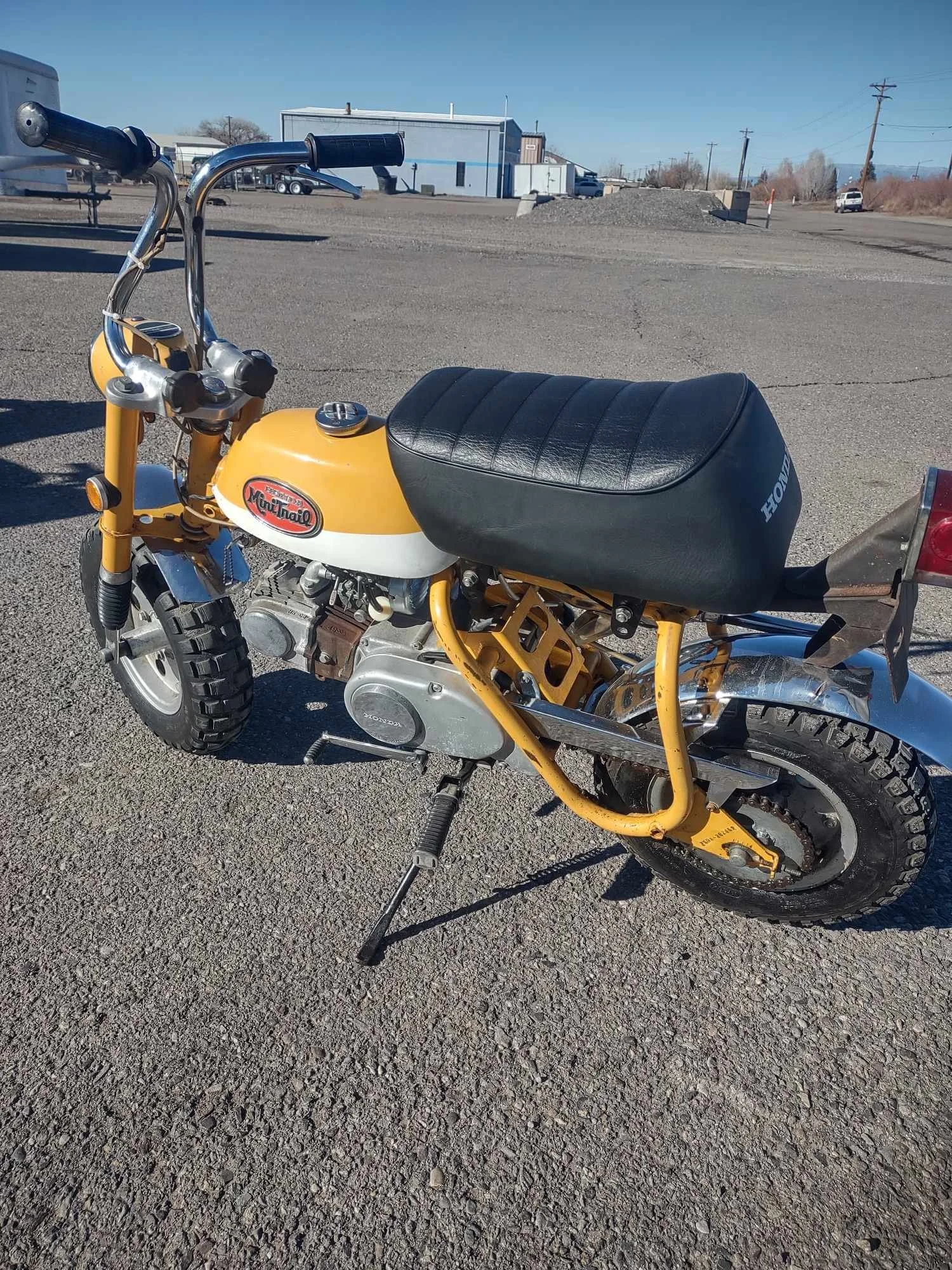 Vintage yellow Honda Monkey mini motorcycle with black seat parked on gravel in an open lot with a few buildings and vehicles in the background.