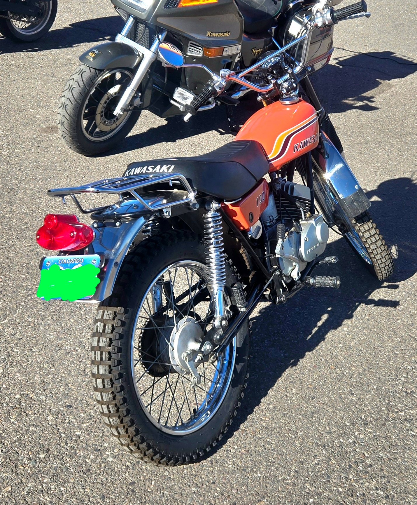 A vintage Kawasaki motorcycle with an orange tank, black seat, and knobby tires, parked on a paved surface with a newer Kawasaki motorcycle in the background.