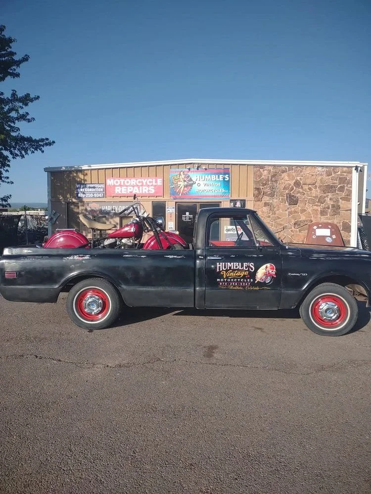 A black pickup truck with red wheels parked in front of a motorbike repair shop. The shop has signs for motorcycle repairs and vintage motorcycle sales, with a vintage red motorcycle and other motorcycle parts in the truck bed.