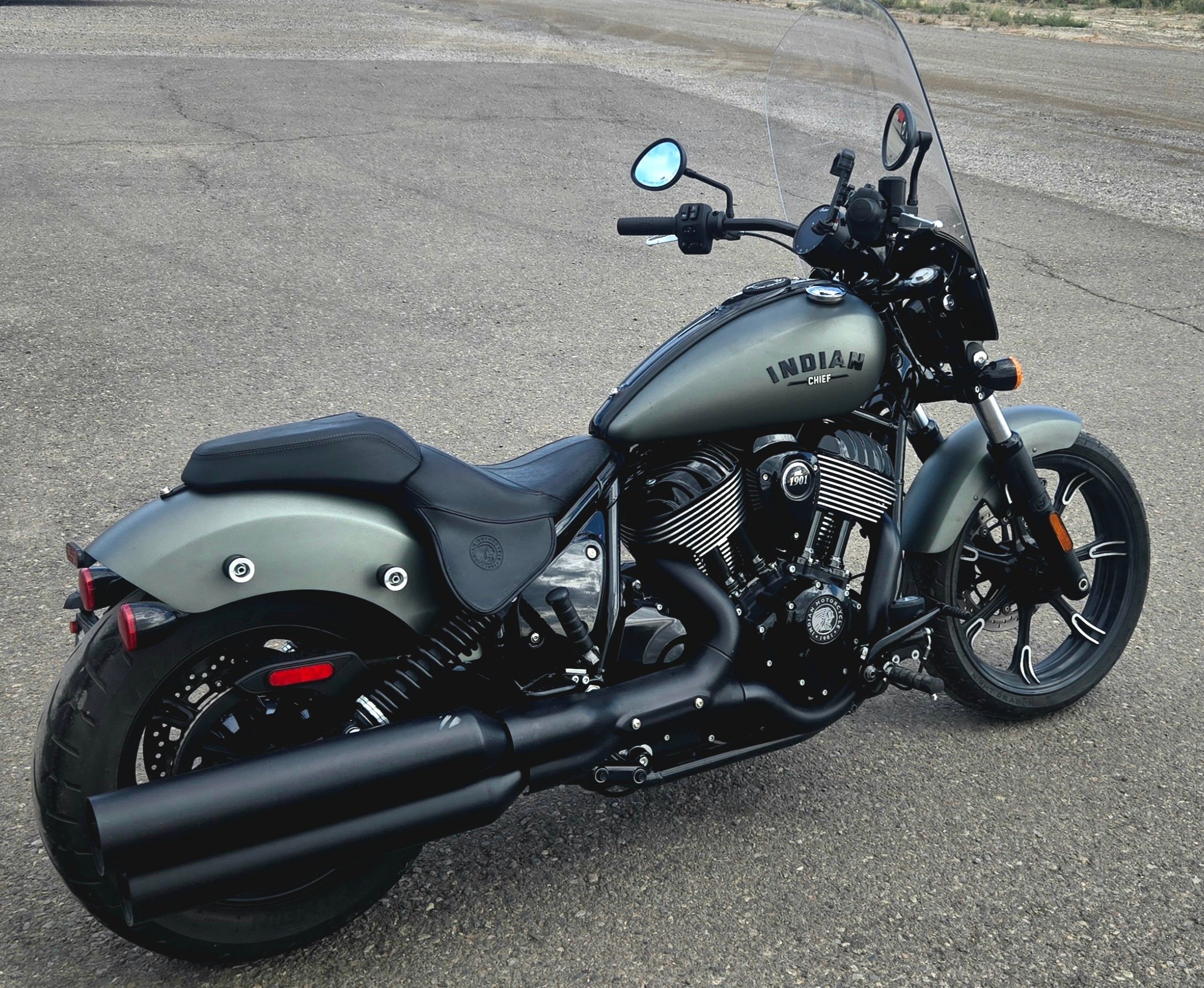 A matte black and gray Indian motorcycle parked on an empty asphalt lot, featuring a windshield, black exhaust, and leather seat.