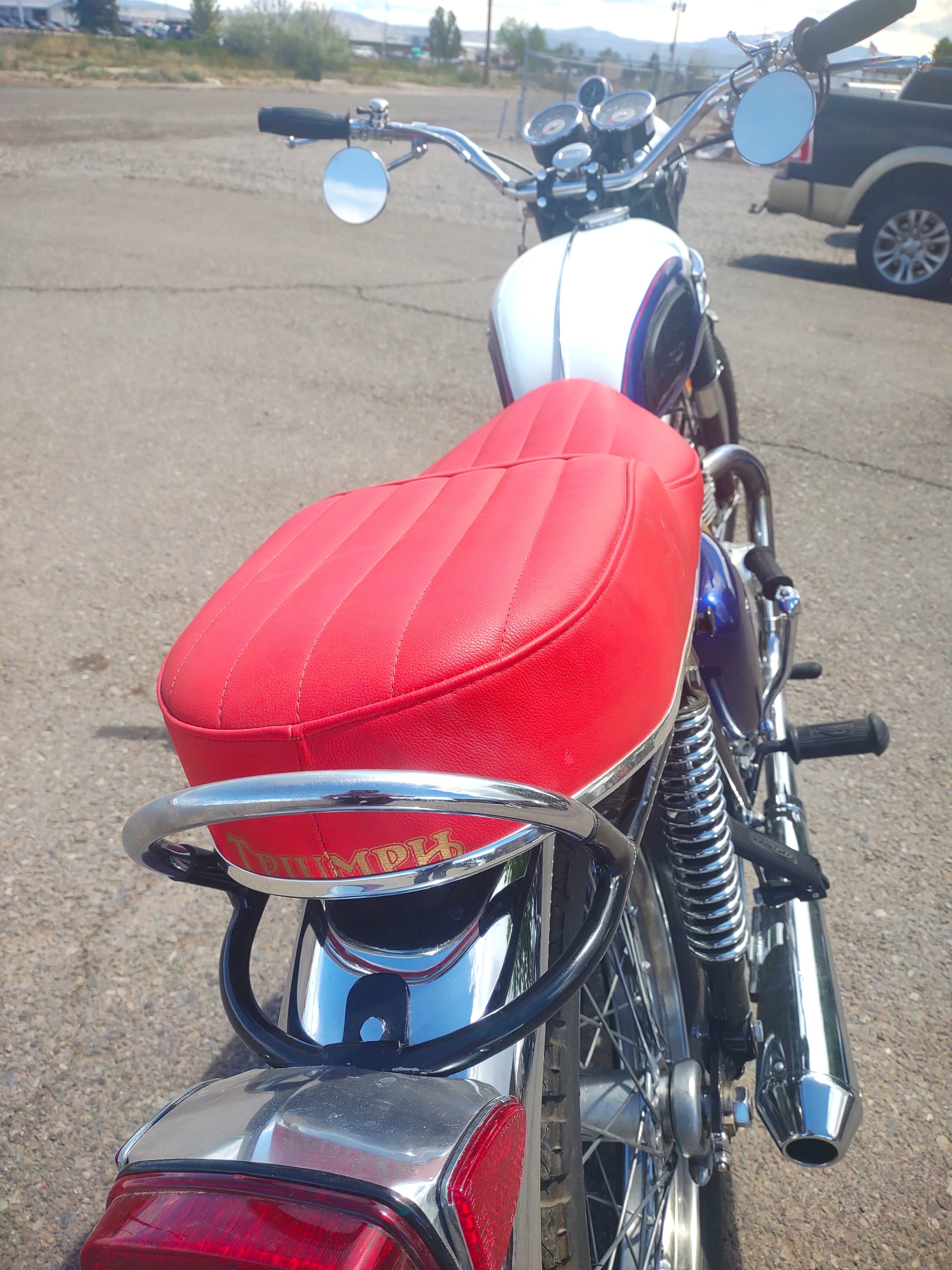 A motorcycle with a red seat and chrome details, parked on a gravel surface, with a white gas tank and black handlebars, and a road with some vehicles and mountains in the background.