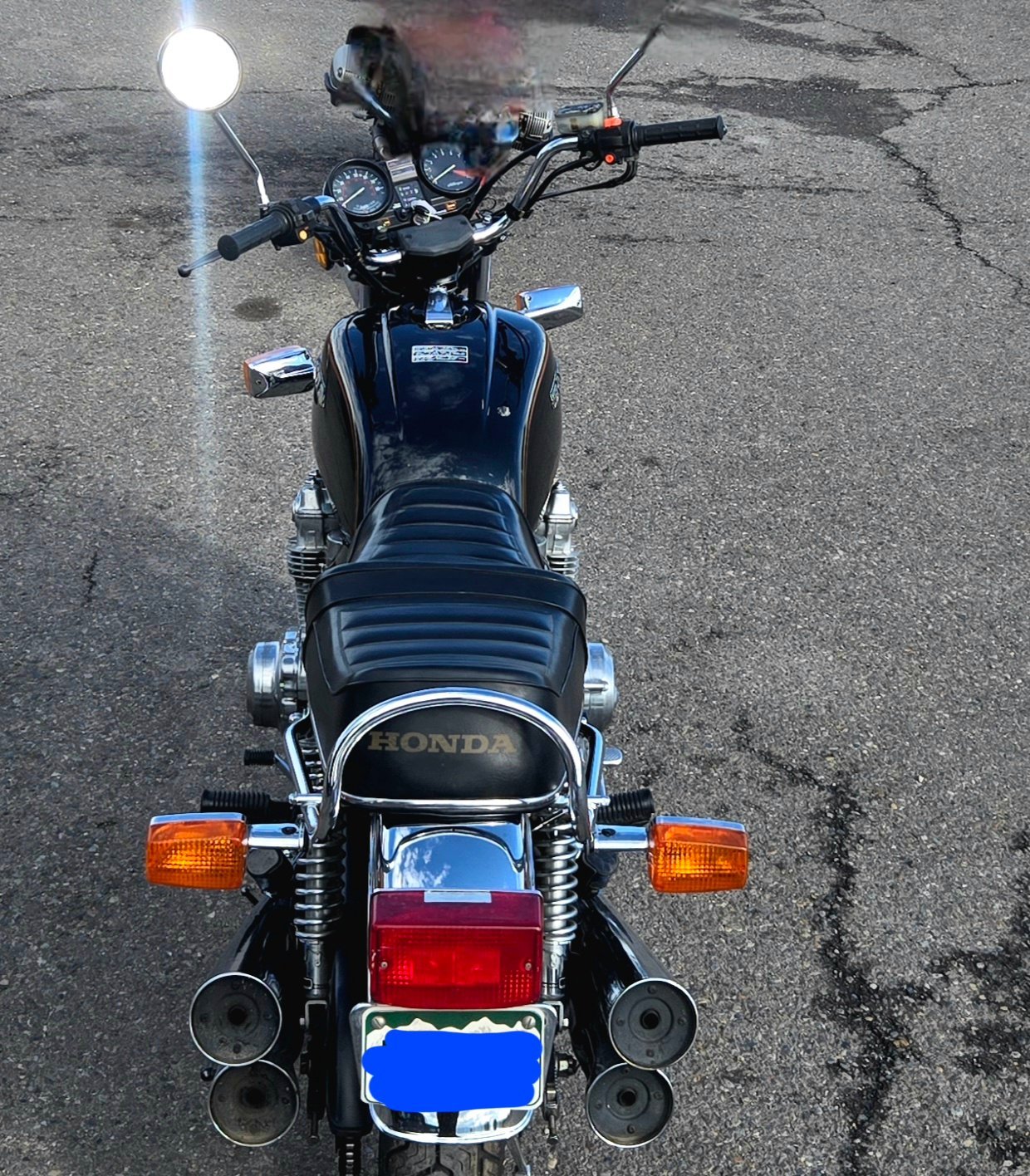 Top view of a black Honda motorcycle parked on asphalt, showing handlebars, seat, rear lights, and exhaust pipes.