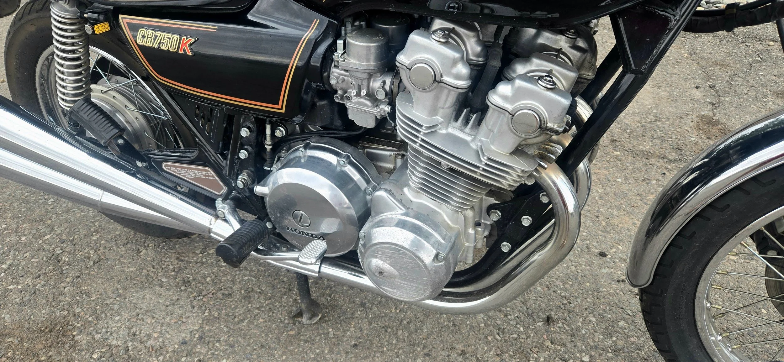 Close-up of a vintage Honda motorcycle engine with black and gold details, chrome exhaust pipes, and black frame, parked on a dirt surface.