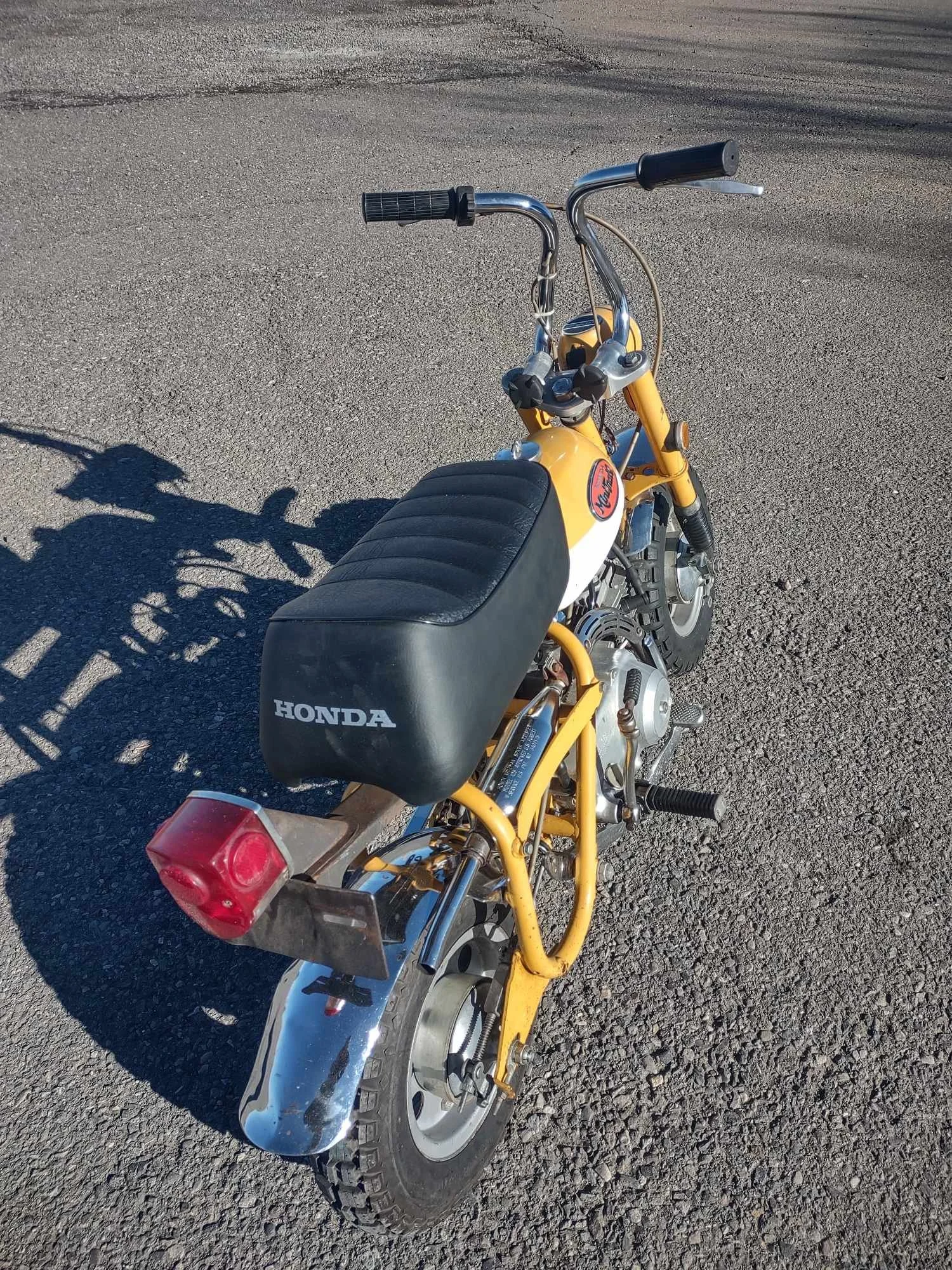 A small yellow Honda mini bike with a black seat and chrome fenders is parked on a gravel road. The handlebars are black with grips, and the bike has a red rear light. The side of the seat displays the Honda logo.