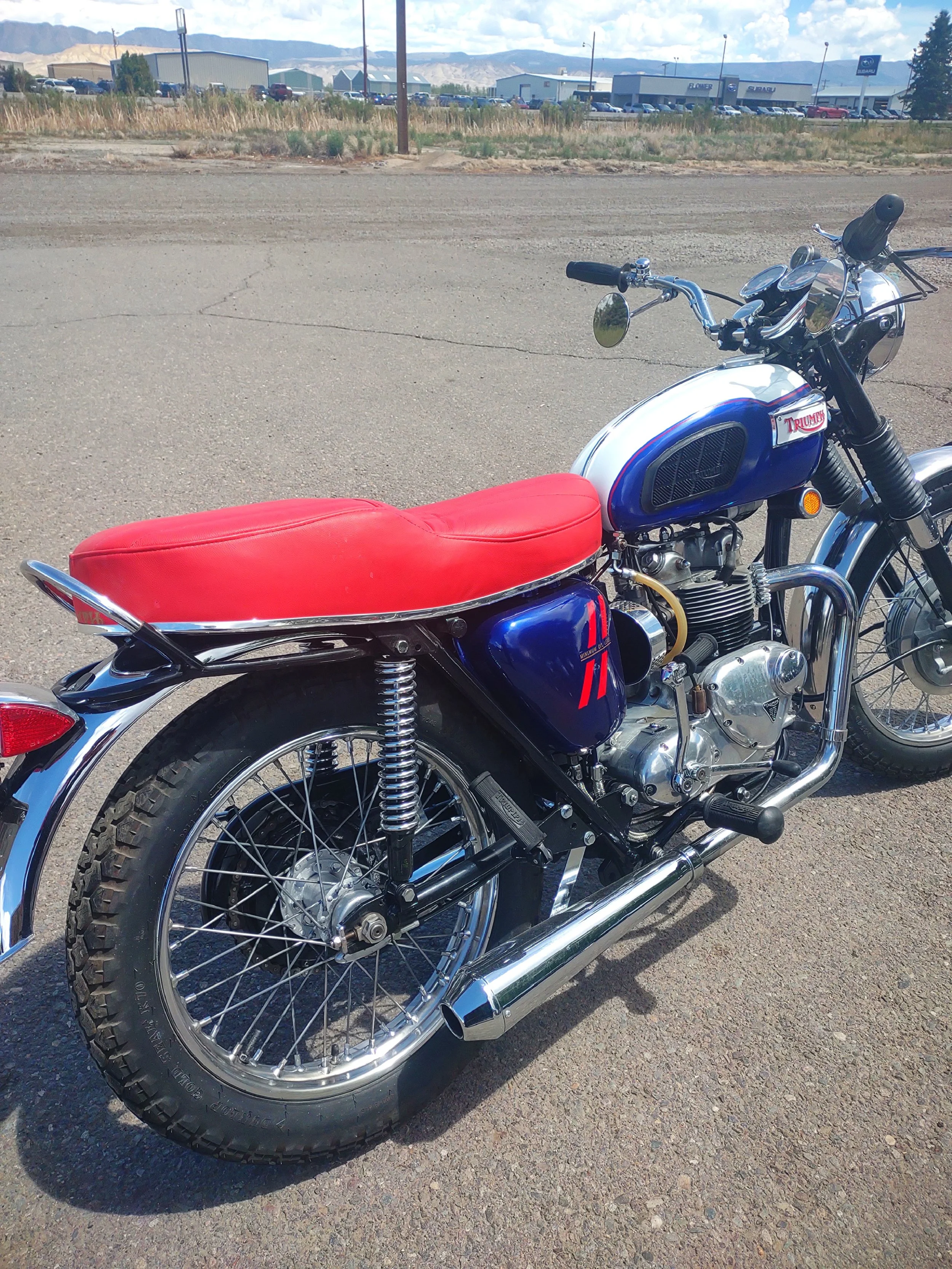 A vintage Triumph motorcycle with a red seat parked on a roadside with industrial buildings and mountains in the background.