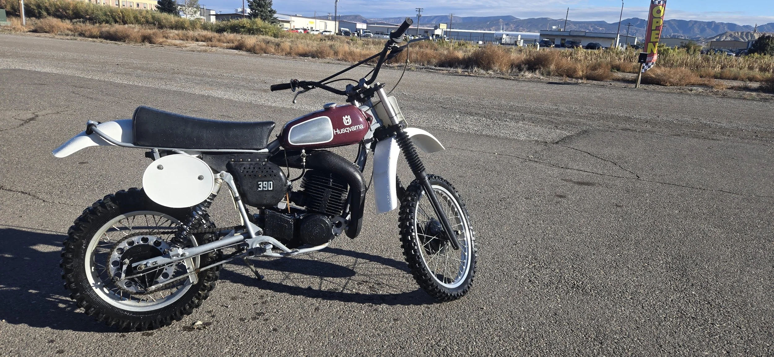 A vintage Husqvarna dirt bike with knobby tires parked on an empty parking lot on a sunny day, with dry grass and mountains in the background.