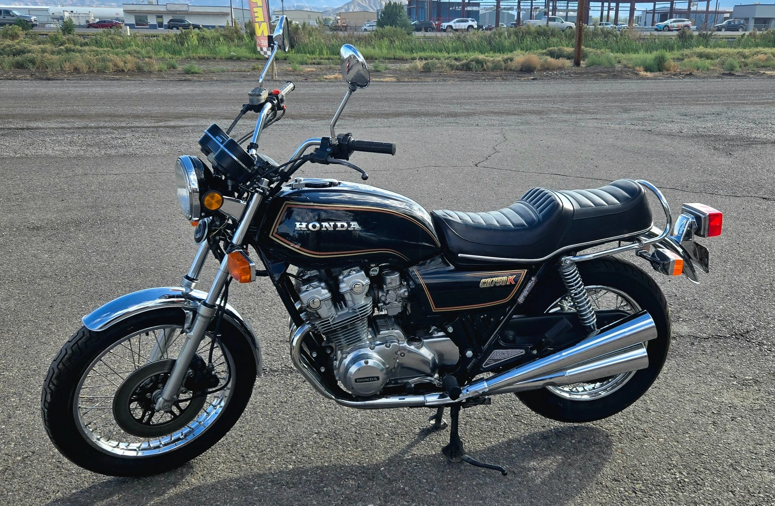 A vintage black Honda CB750K motorcycle parked on a gravel lot with a building and cars in the background.