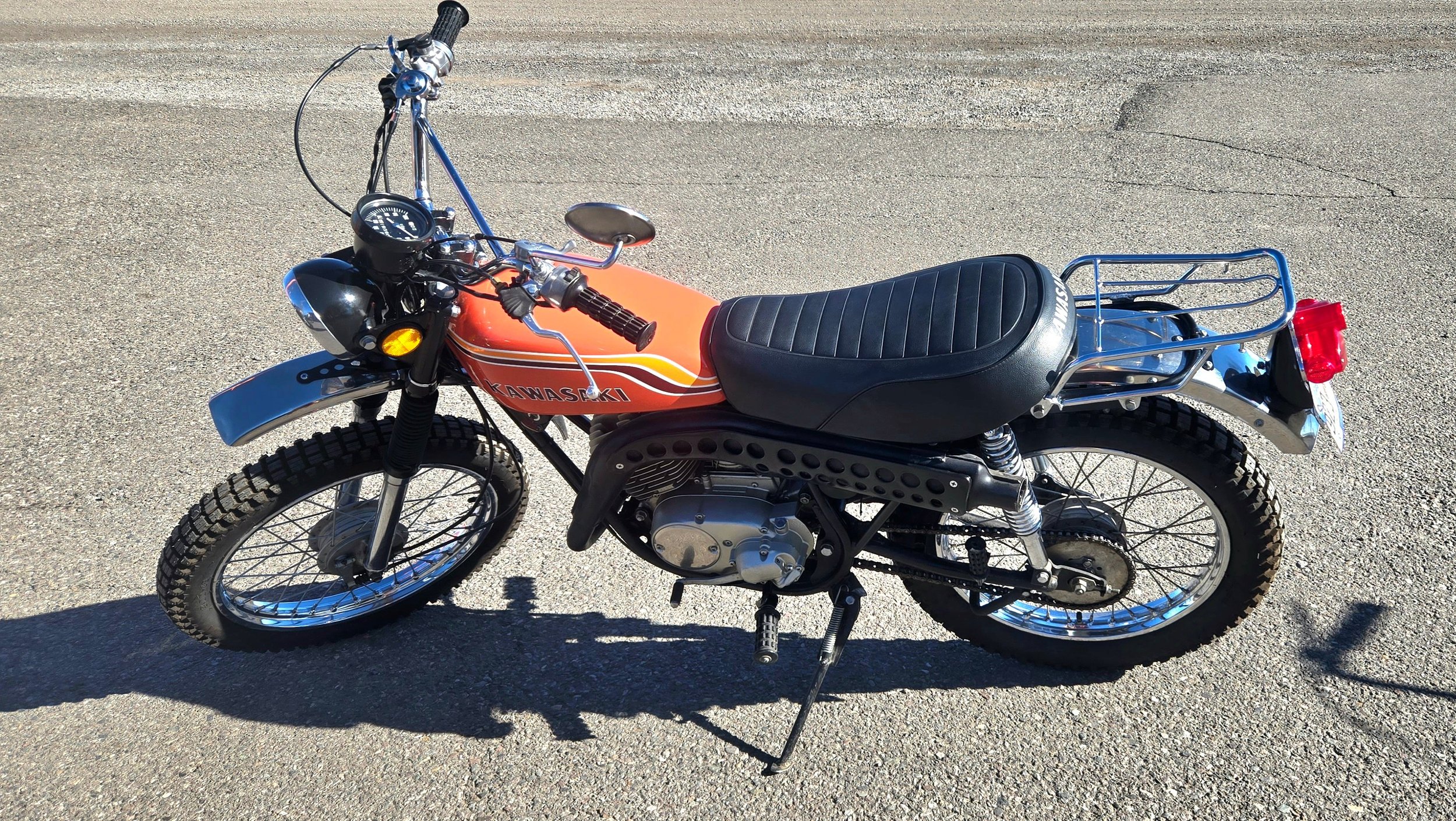 A vintage Kawasaki dirt bike with a black seat, orange fuel tank, and chrome accents, parked on asphalt.