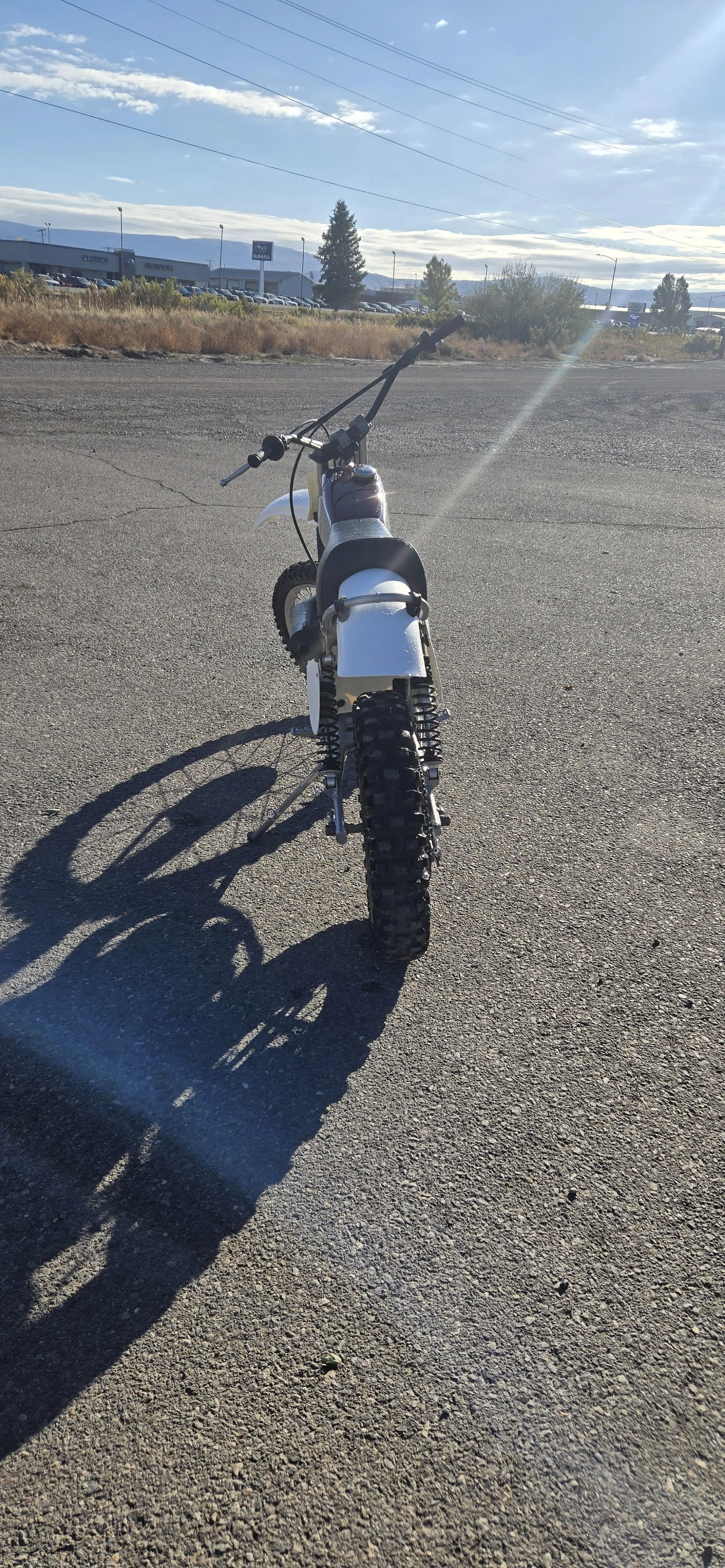 Front view of a small dirt bike on a gravel surface with a dealership and parking lot in the background under a partly cloudy sky.