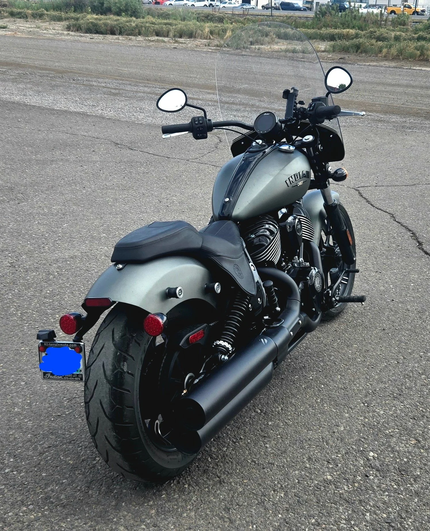 Black and matte gray Indian motorcycle parked on an asphalt surface with a background of fields, trees, and a parking lot with cars.