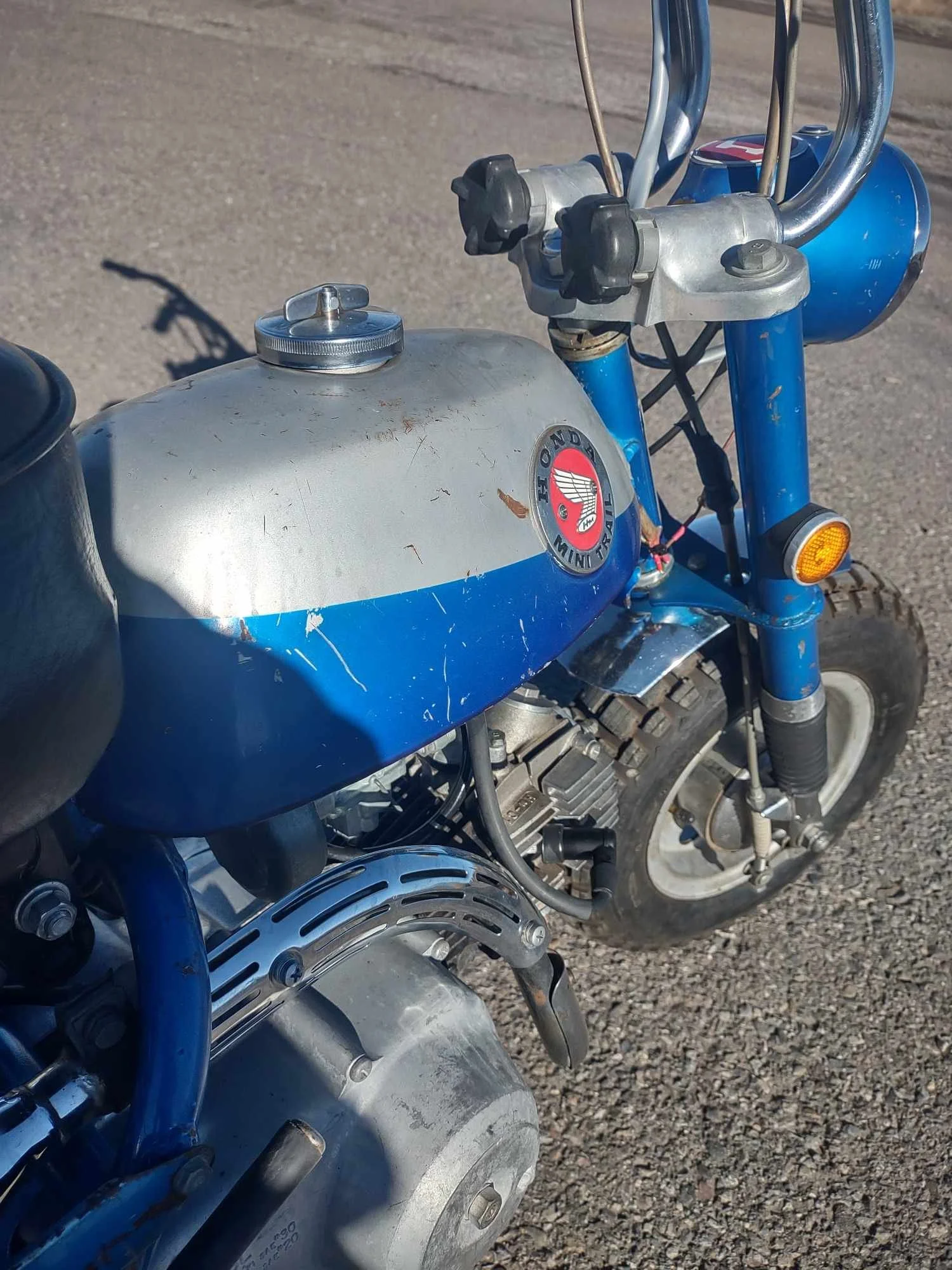 Close-up of a vintage Honda Mini Trail motorcycle with a blue and silver fuel tank, showing signs of wear and rust, set on a gravel surface.