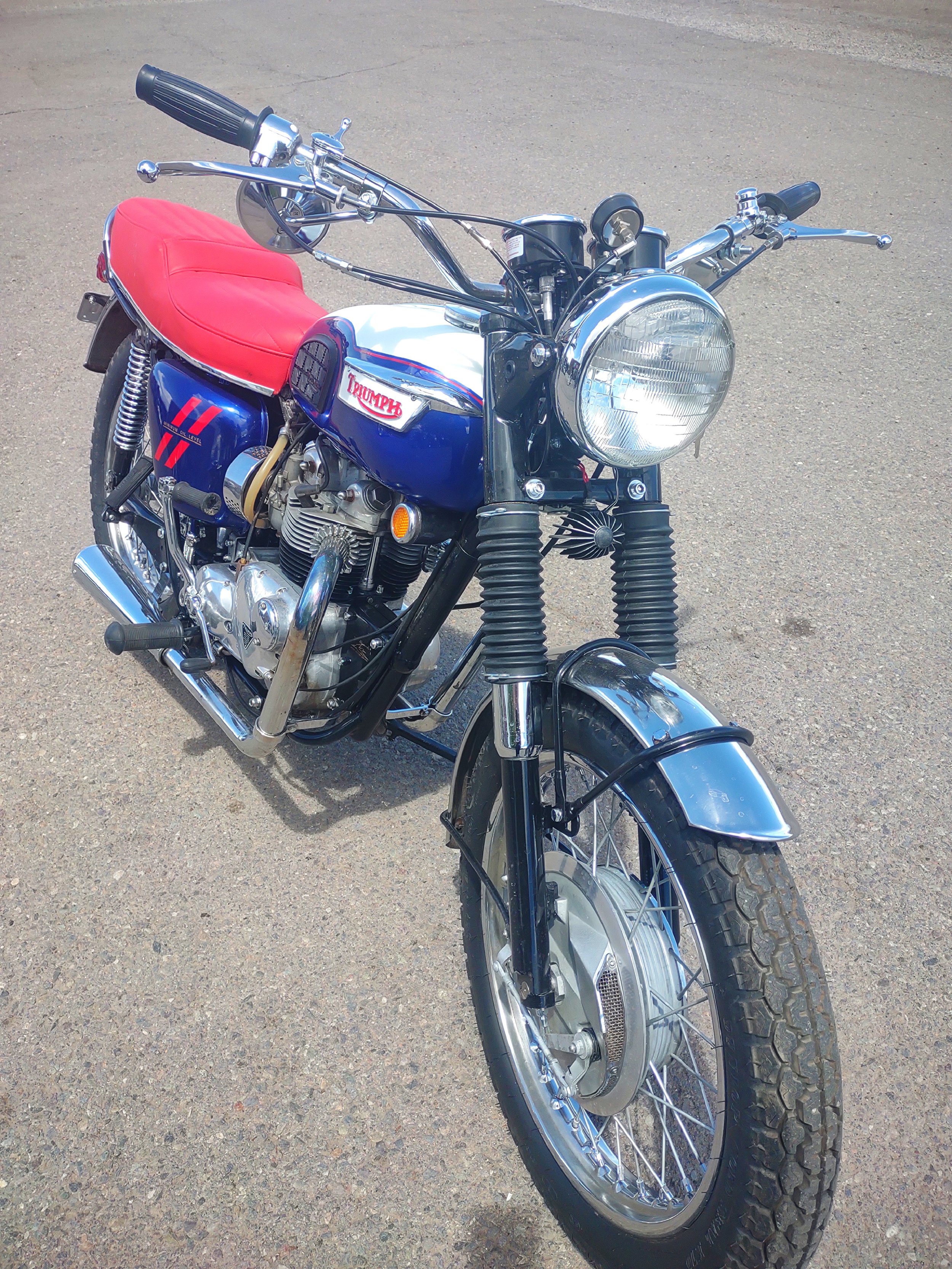 A vintage blue Triumph motorcycle with a red seat, chrome accents, and an electric front motor, parked on asphalt.