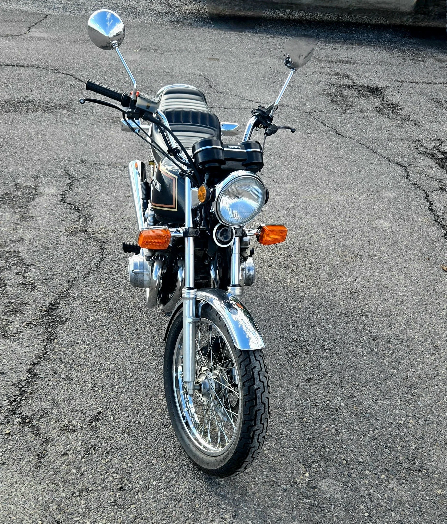 Front view of a vintage black and chrome motorcycle with a round headlight, handlebar mirrors, and a padded seat on a cracked asphalt surface.