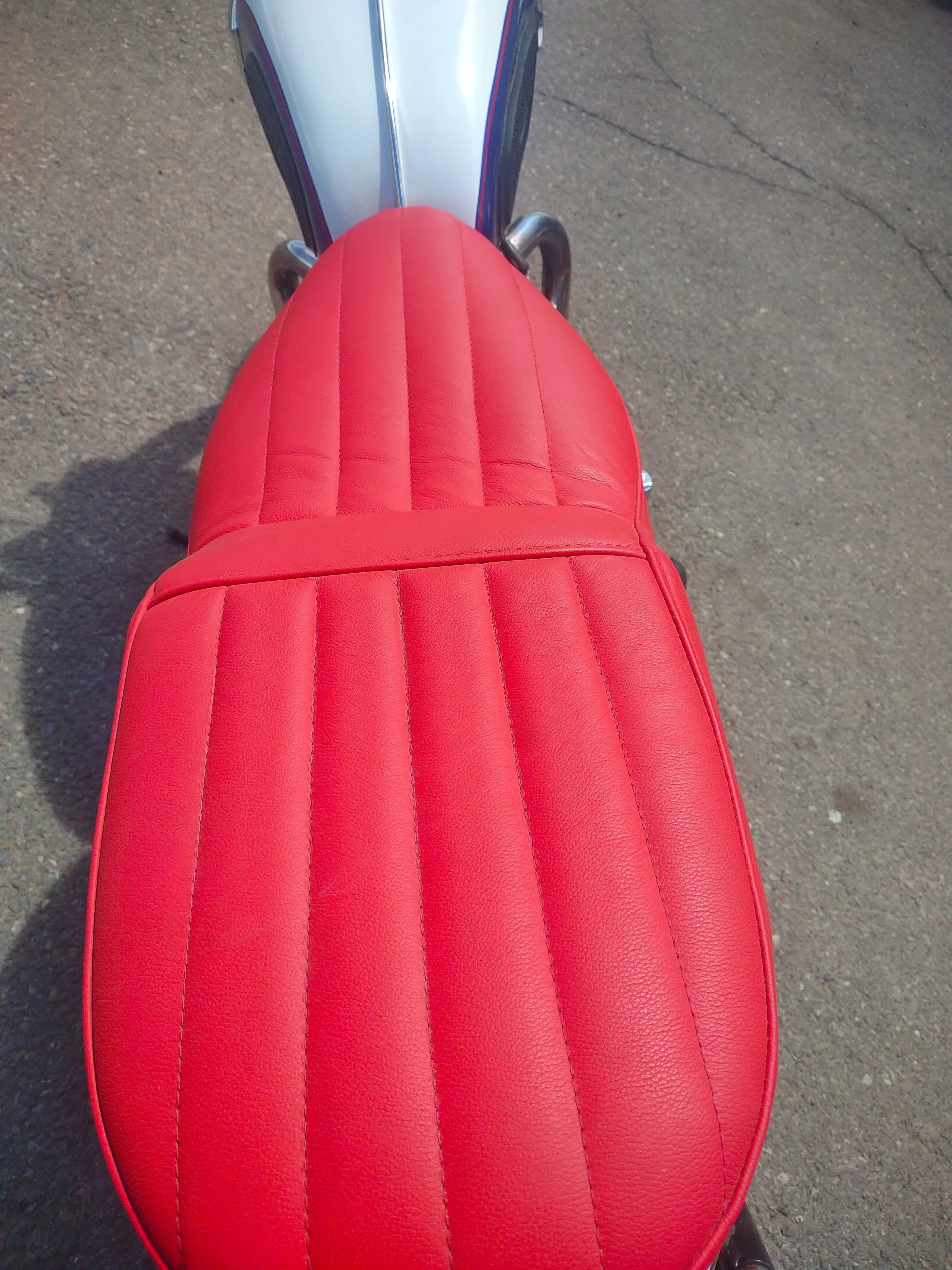Top view of a red seat on a motorcycle with a silver and blue fuel tank visible in the background, parked on asphalt.