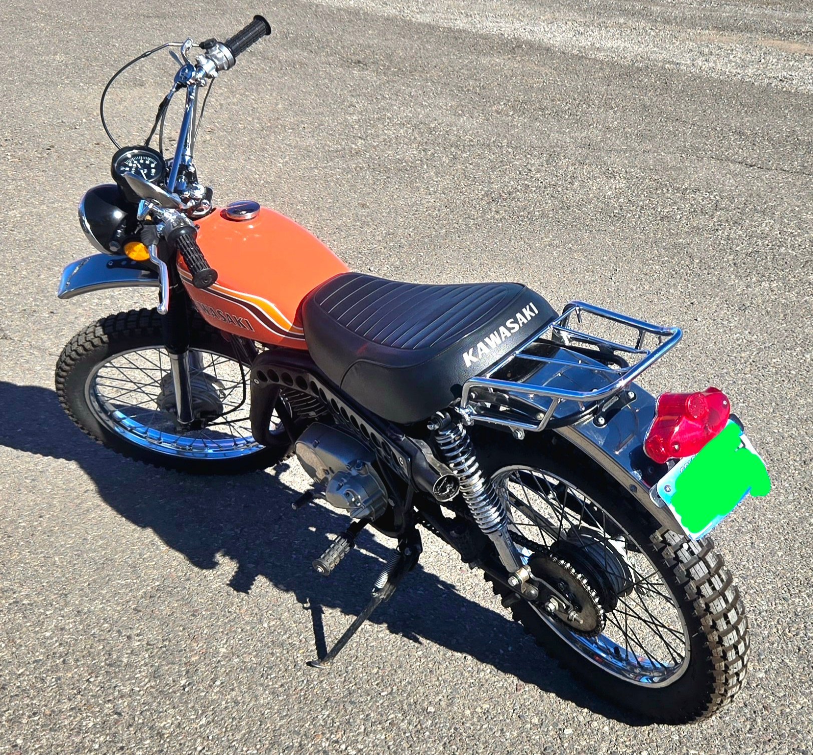 A vintage Kawasaki dirt bike with an orange fuel tank, black seat, knobby tires, and chrome accents parked on a paved surface.