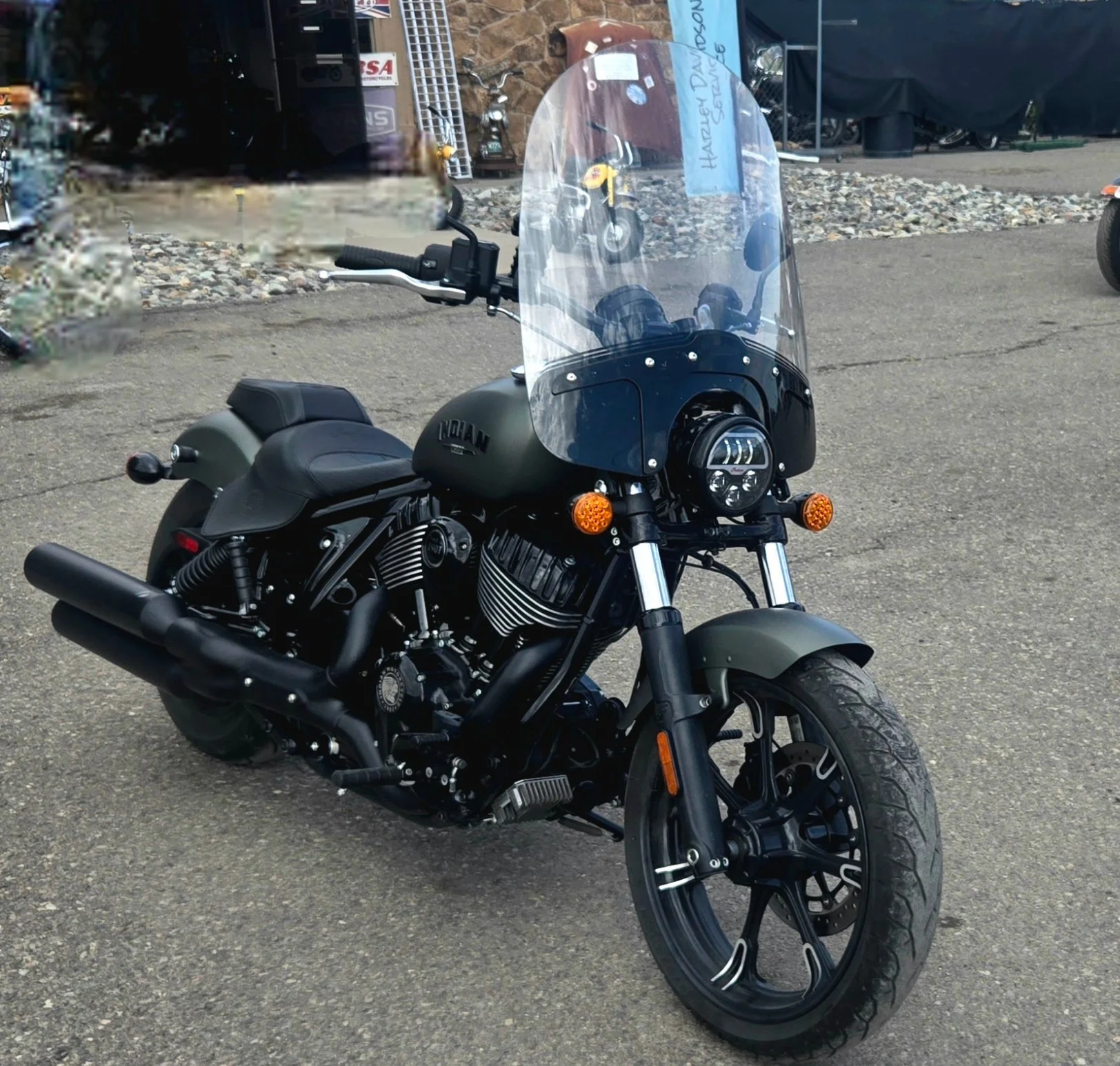 Black cruiser motorcycle with a windshield parked on pavement, with a backdrop of gravel and buildings.
