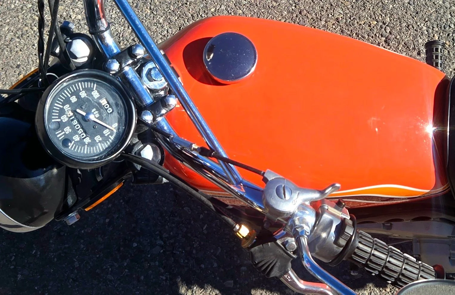 Top view of a vintage orange motorcycle's dashboard and fuel tank, with a speedometer, handlebars, and black grips visible.