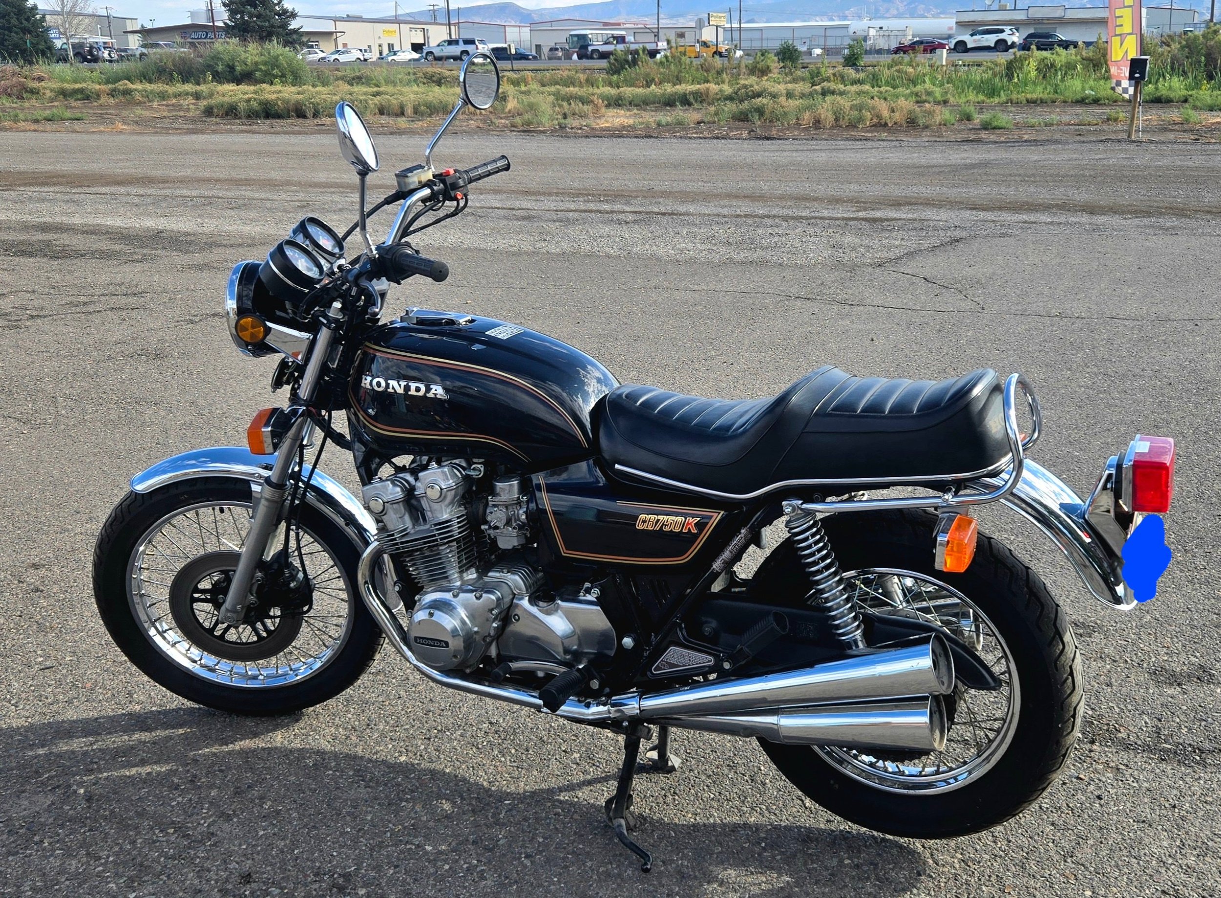 A black vintage Honda CB750K motorcycle parked on a paved lot with a background of stores, vehicles, and a mountain range under a clear sky.