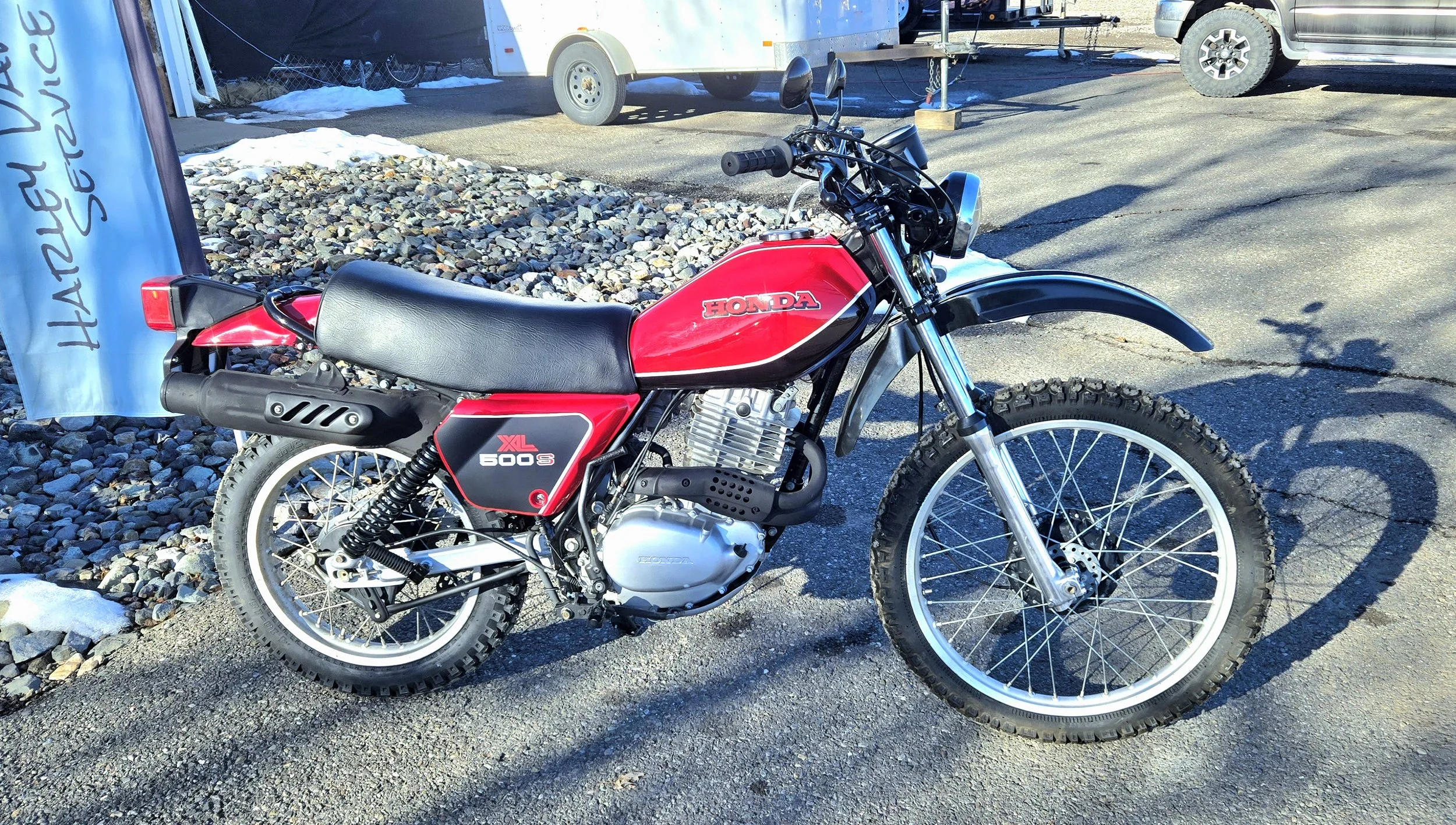 A vintage Honda XL 600 S off-road motorcycle parked on a pavement with gravel and snow nearby, with a trailer and vehicles in the background during daytime.