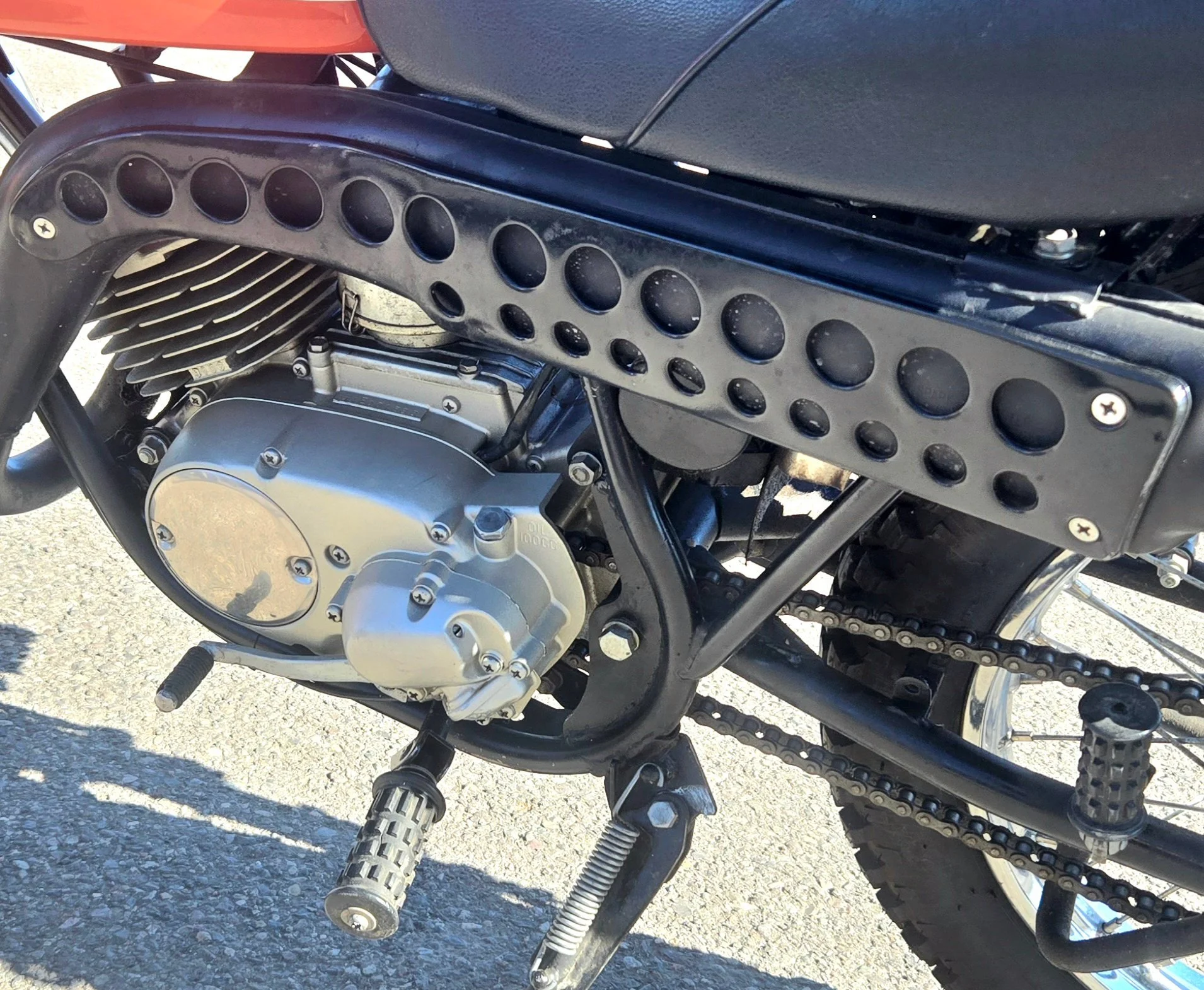Close-up of a motorcycle's engine, chain, and footpeg, with a black perforated footguard and a black frame, on a paved road.