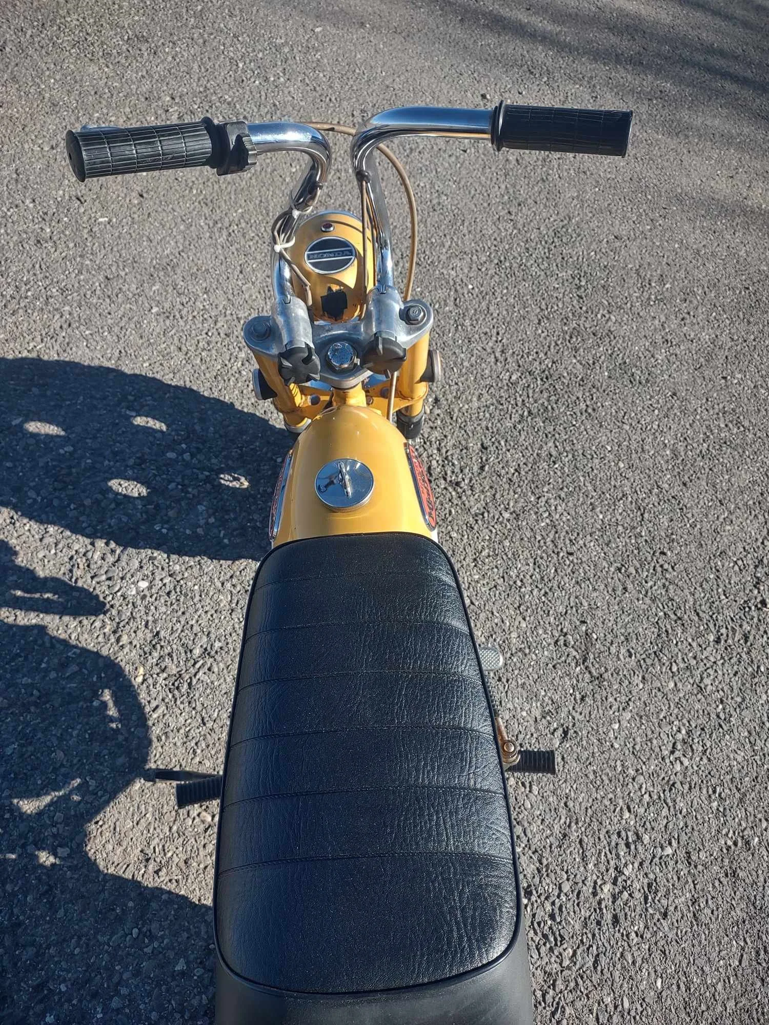 Top-down view of a yellow vintage motorcycle with black seat, chrome handlebars, and gauges, parked on a gravel surface.