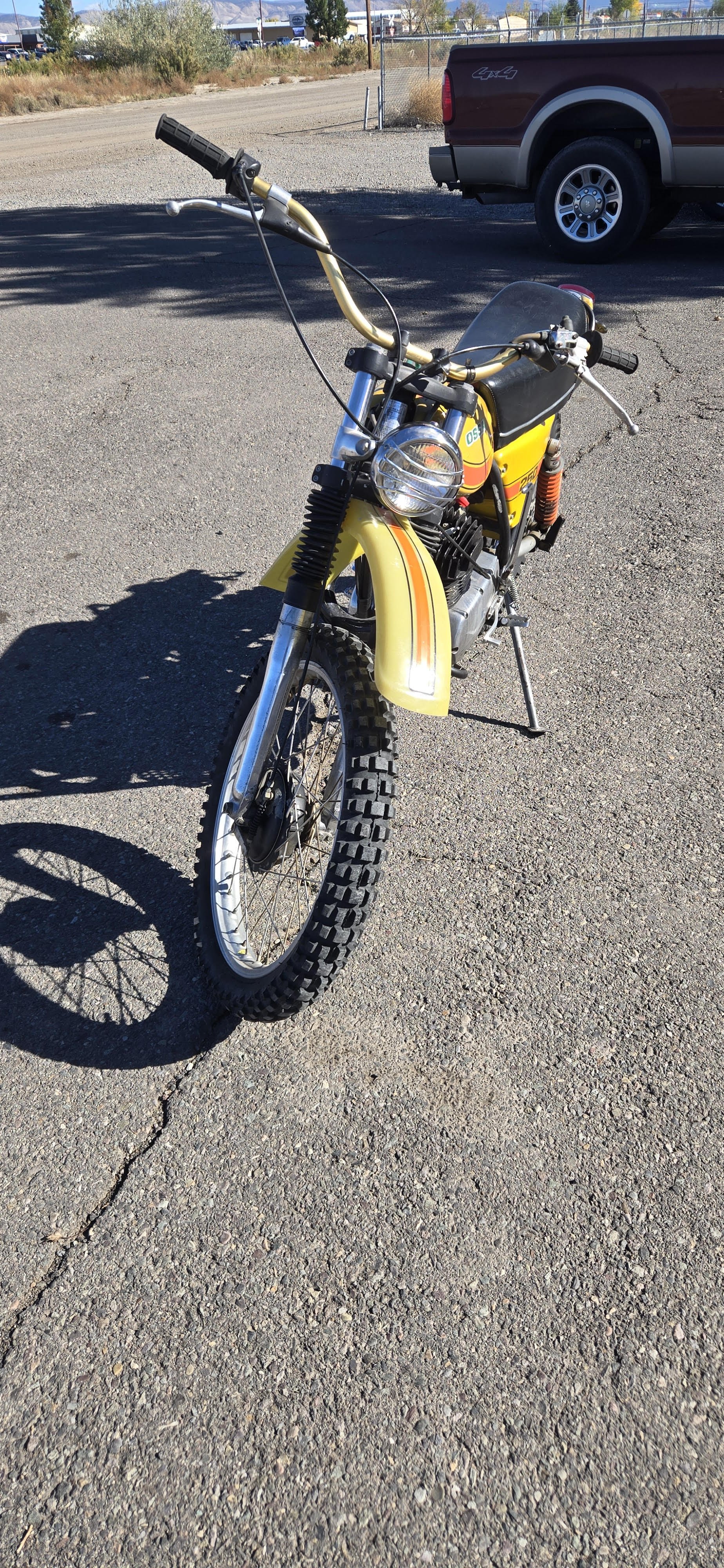 Vintage yellow dirt bike with black seat and knobby tires, parked on a gravel lot with a red pickup truck in the background.