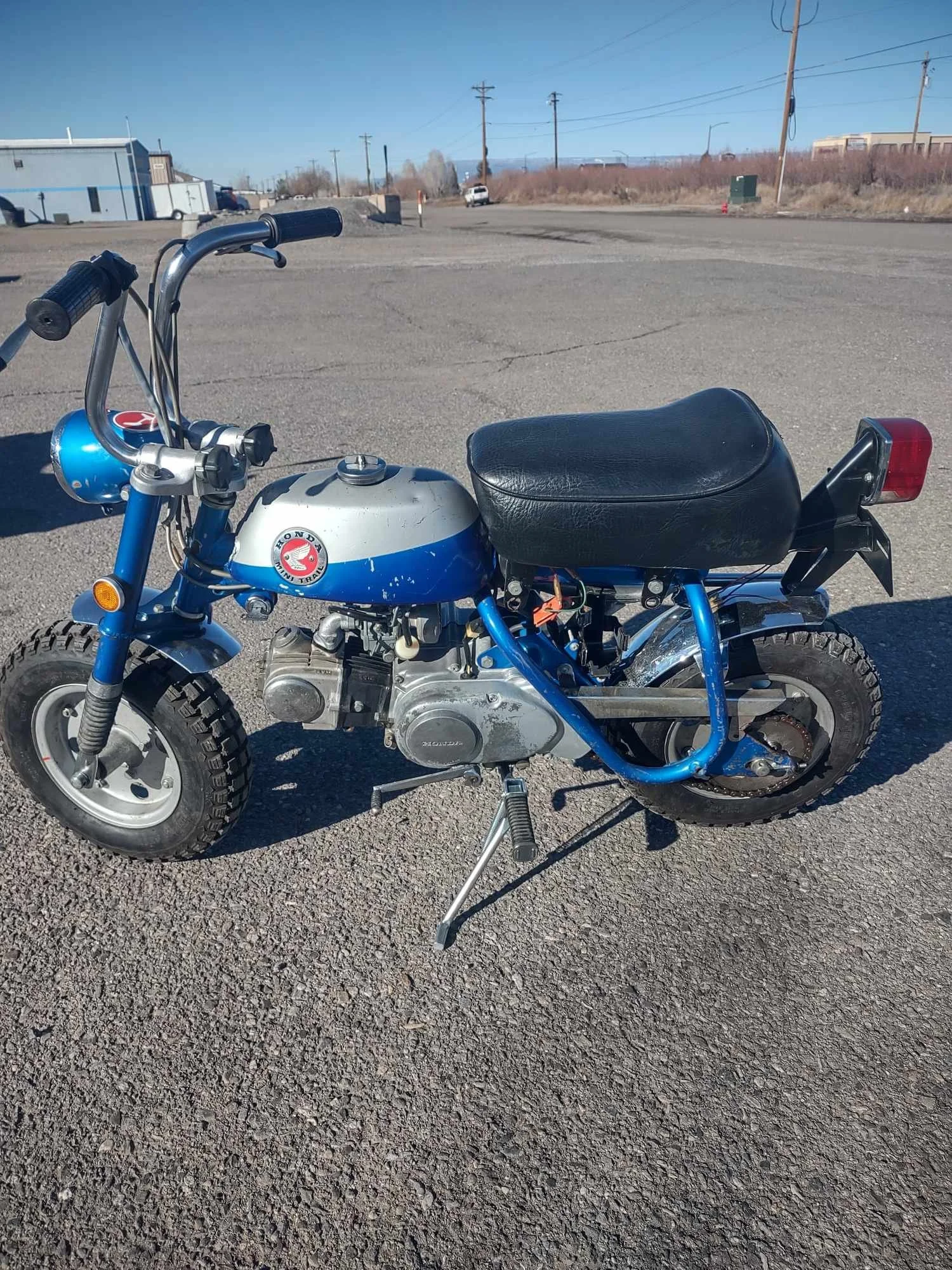 A vintage Honda mini bike with a blue and silver fuel tank, black seat, and knobby tires, parked on an empty asphalt lot with utility poles and industrial buildings in the background.