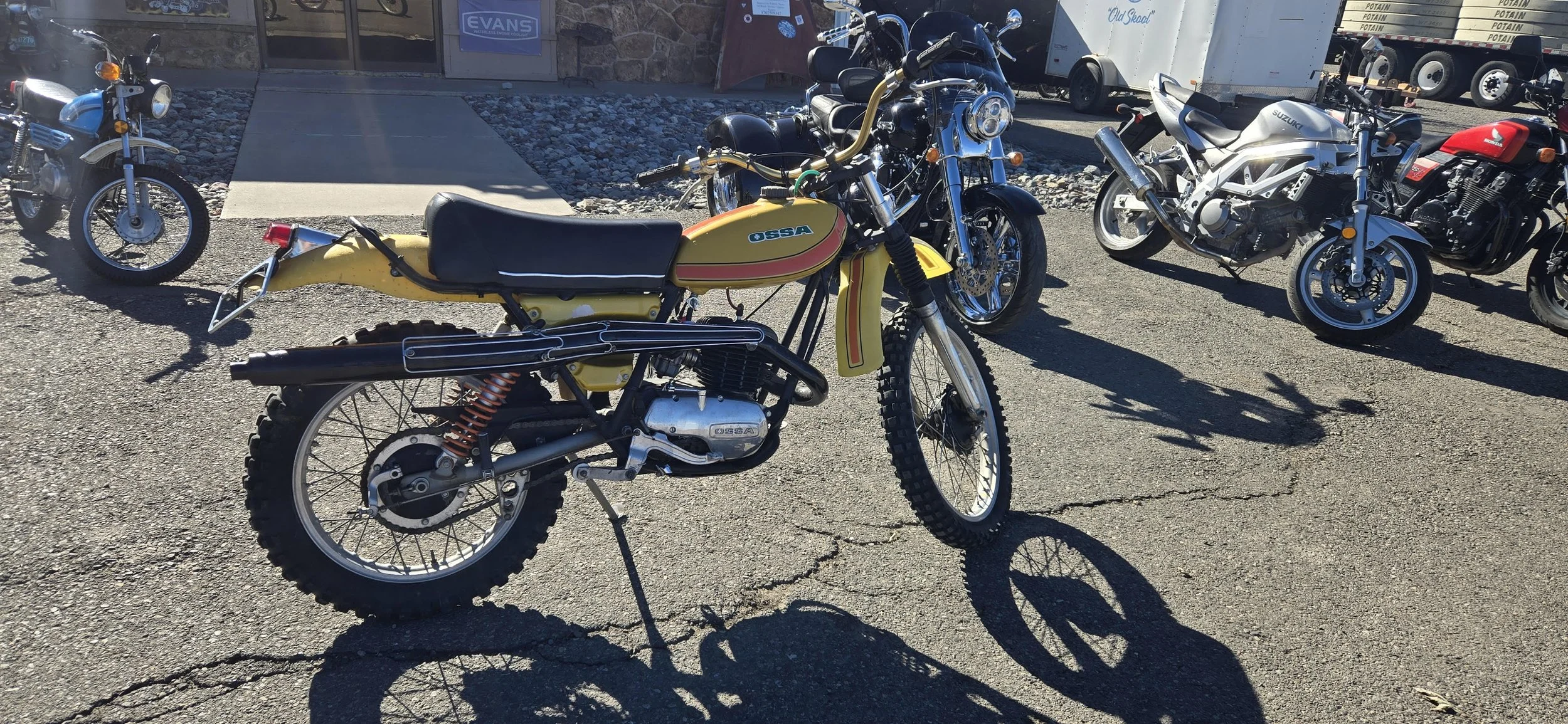 A yellow vintage OSSA dirt bike with off-road tires parked on a cracked asphalt surface in front of a building, with several motorcycles and a trailer in the background.