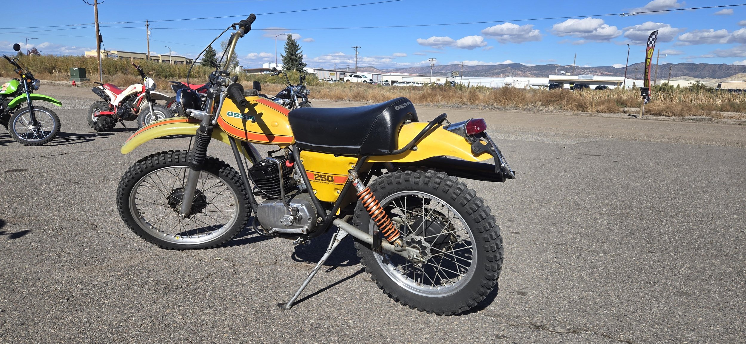 Yellow vintage dirt bike with knobby tires, parked on a gravel lot, with other bikes in the background and mountains in the distance under a partly cloudy sky.
