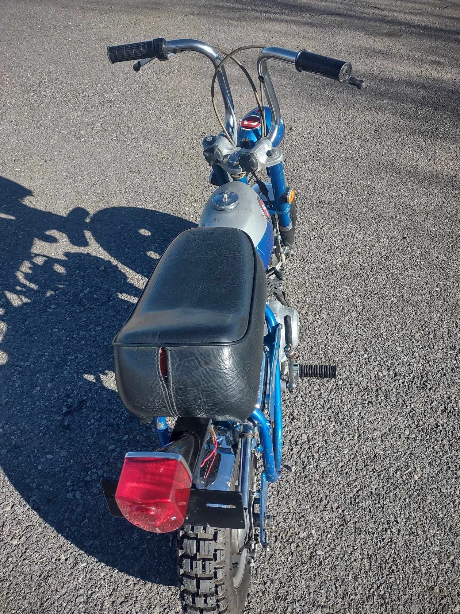 A small blue motorcycle with a black seat, black handlebars, and a red rear light parked on a gravel surface.