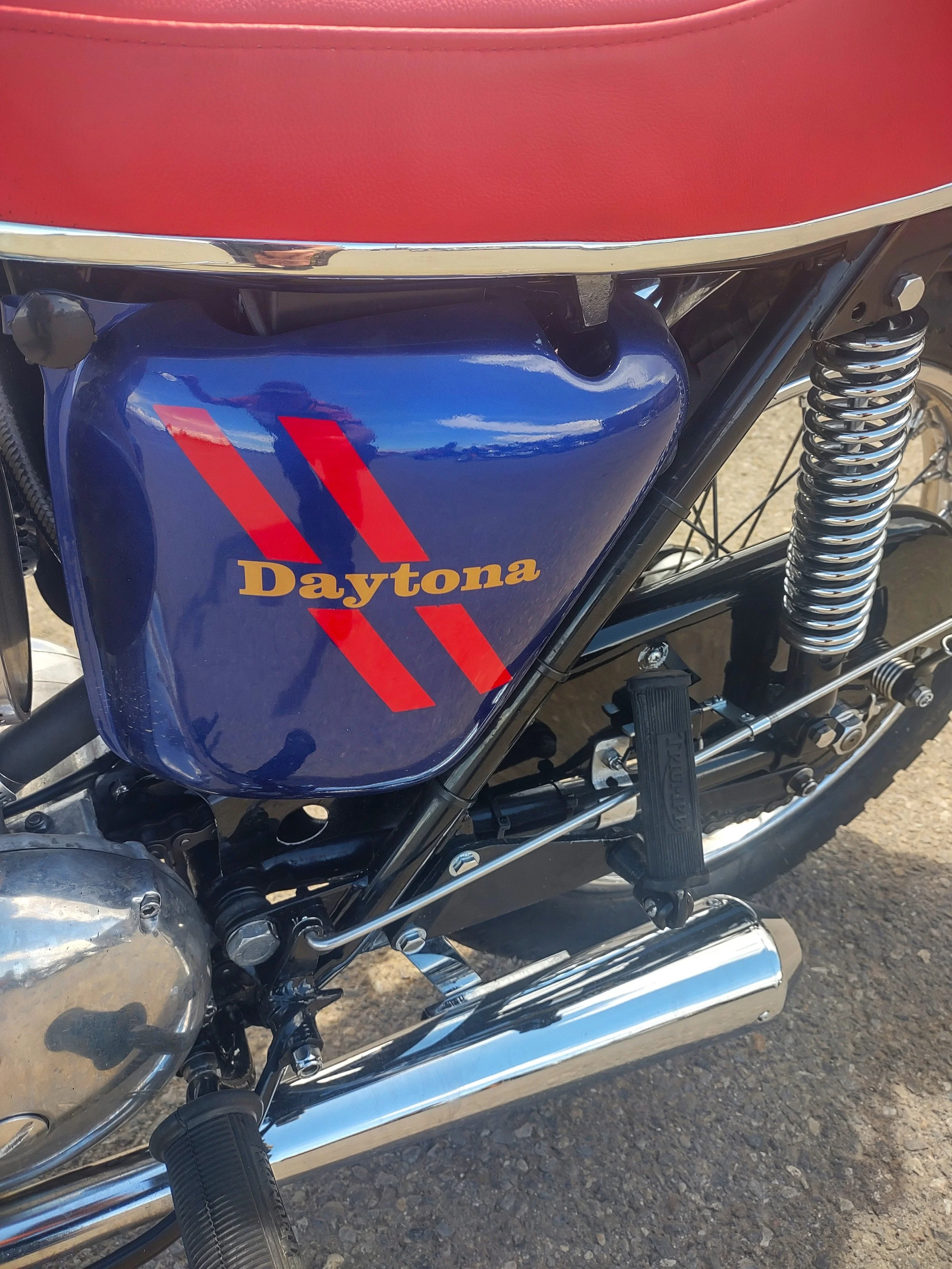 Close-up of a blue motorcycle fuel tank with red stripes and the word 'Daytona' written in yellow, along with parts of the engine, foot pegs, and rear suspension.