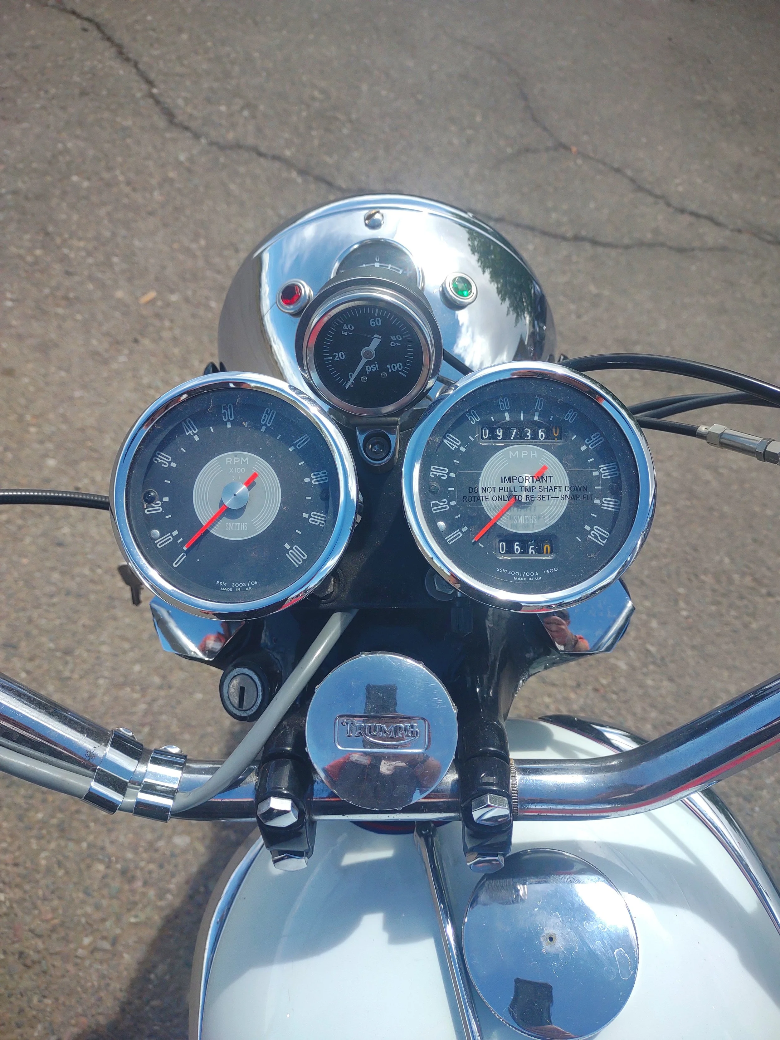 View of a Triumph motorcycle's dashboard with speedometer, tachometer, and other gauges, on a paved surface.