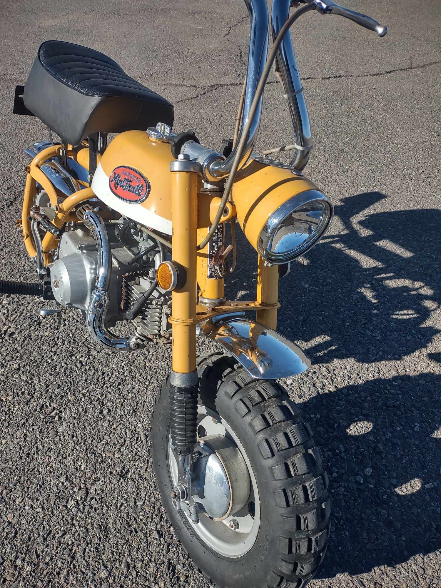 A vintage yellow Honda MotraMotor mini motorcycle with a black seat, chrome handlebars, and large off-road front tires, parked on asphalt.