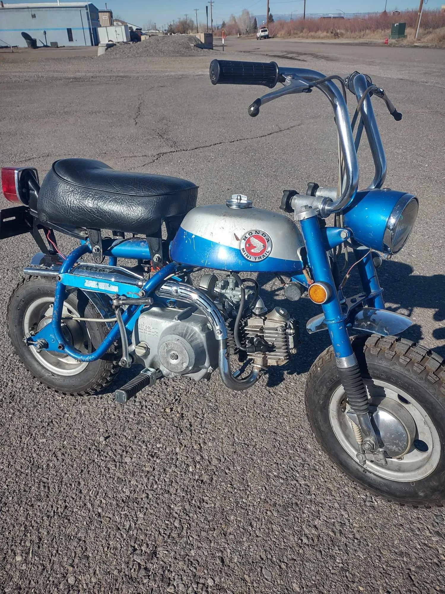 A small vintage Honda mini bike with a blue frame, silver gas tank, and black seat, parked on a gravel lot.