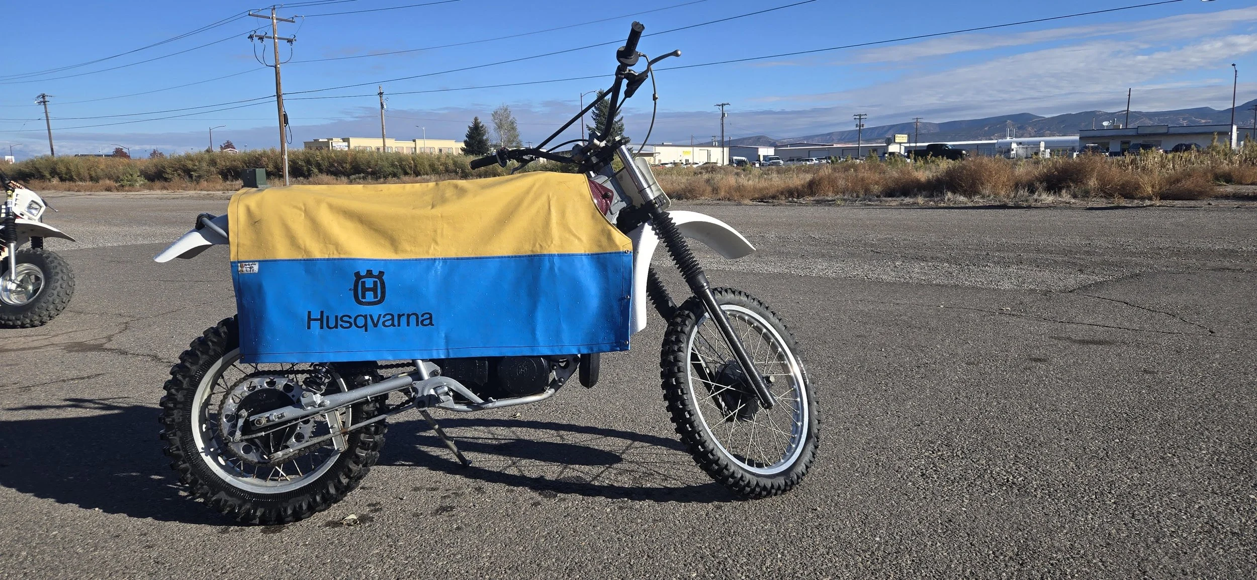 A Husqvarna electric dirt bike with a yellow and blue cover parked on an asphalt lot with another dirt bike partially visible to the left. Industrial buildings, power lines, and mountains are in the background under a partly cloudy sky.